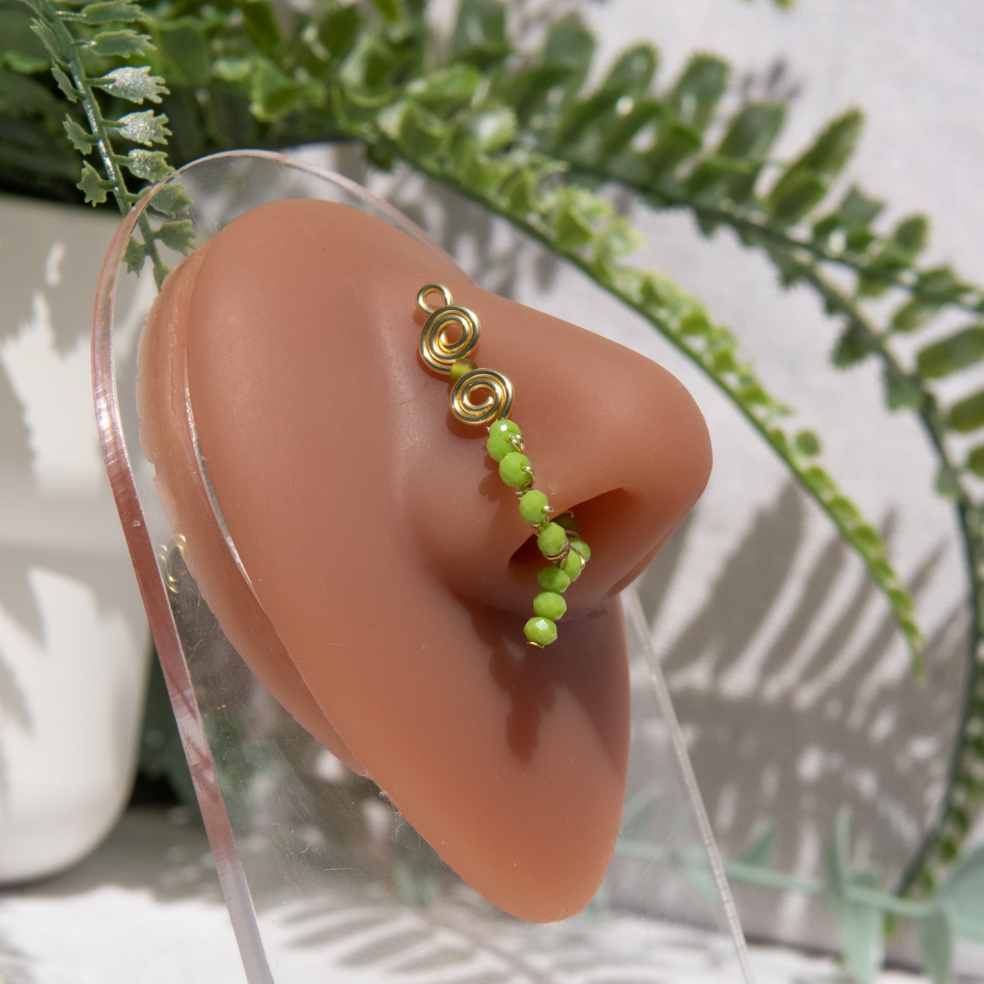 A light gold-toned faux dangle nose ring cuffs a model's nose. Bright green seed beads are wire-wrapped around the hoop's circumference, with a beaded wire dangling down.