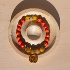 A beaded stretch bracelet with orange/yellow marbled beads, Old Topaz stone beads, and a gold-plated orange enamel palm tree circle charm, displayed on a circular wooden stand against a light wood background