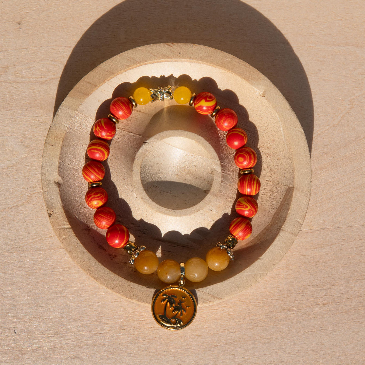 A beaded stretch bracelet with orange/yellow marbled beads, Old Topaz stone beads, and a gold-plated orange enamel palm tree circle charm, displayed on a circular wooden stand against a light wood background