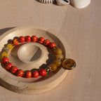 A beaded stretch bracelet featuring orange/yellow marbled beads, Old Topaz stone beads, and a gold-plated orange enamel palm tree circle charm, displayed on a circular wooden stand against a light wood background.