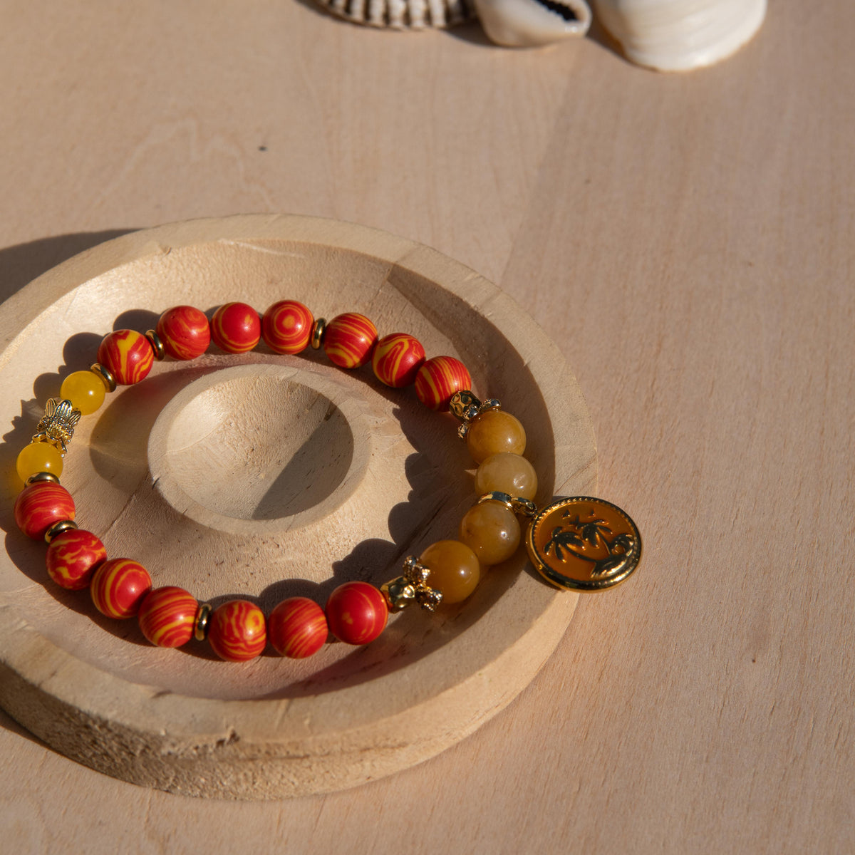 A beaded stretch bracelet featuring orange/yellow marbled beads, Old Topaz stone beads, and a gold-plated orange enamel palm tree circle charm, displayed on a circular wooden stand against a light wood background.