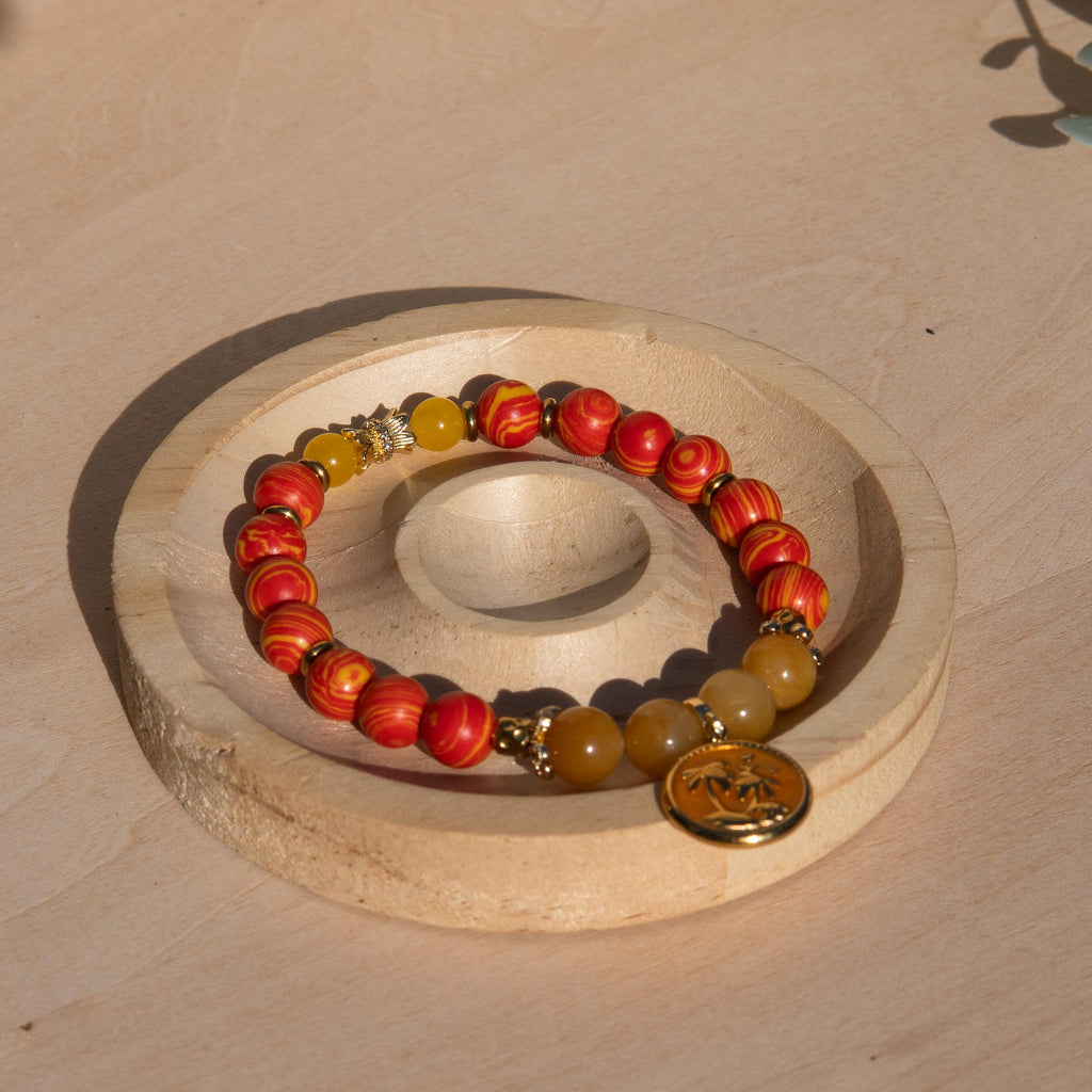 A beaded stretch bracelet featuring orange/yellow marbled beads, Old Topaz stone beads, and a gold-plated orange enamel palm tree circle charm, displayed on a circular wooden stand against a light wood background.
