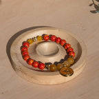 A beaded stretch bracelet featuring orange/yellow marbled beads, Old Topaz stone beads, and a gold-plated orange enamel palm tree circle charm, displayed on a circular wooden stand against a light wood background.