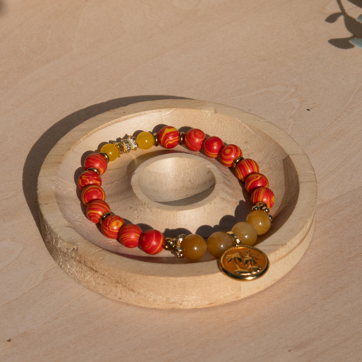A beaded stretch bracelet featuring orange/yellow marbled beads, Old Topaz stone beads, and a gold-plated orange enamel palm tree circle charm, displayed on a circular wooden stand against a light wood background.