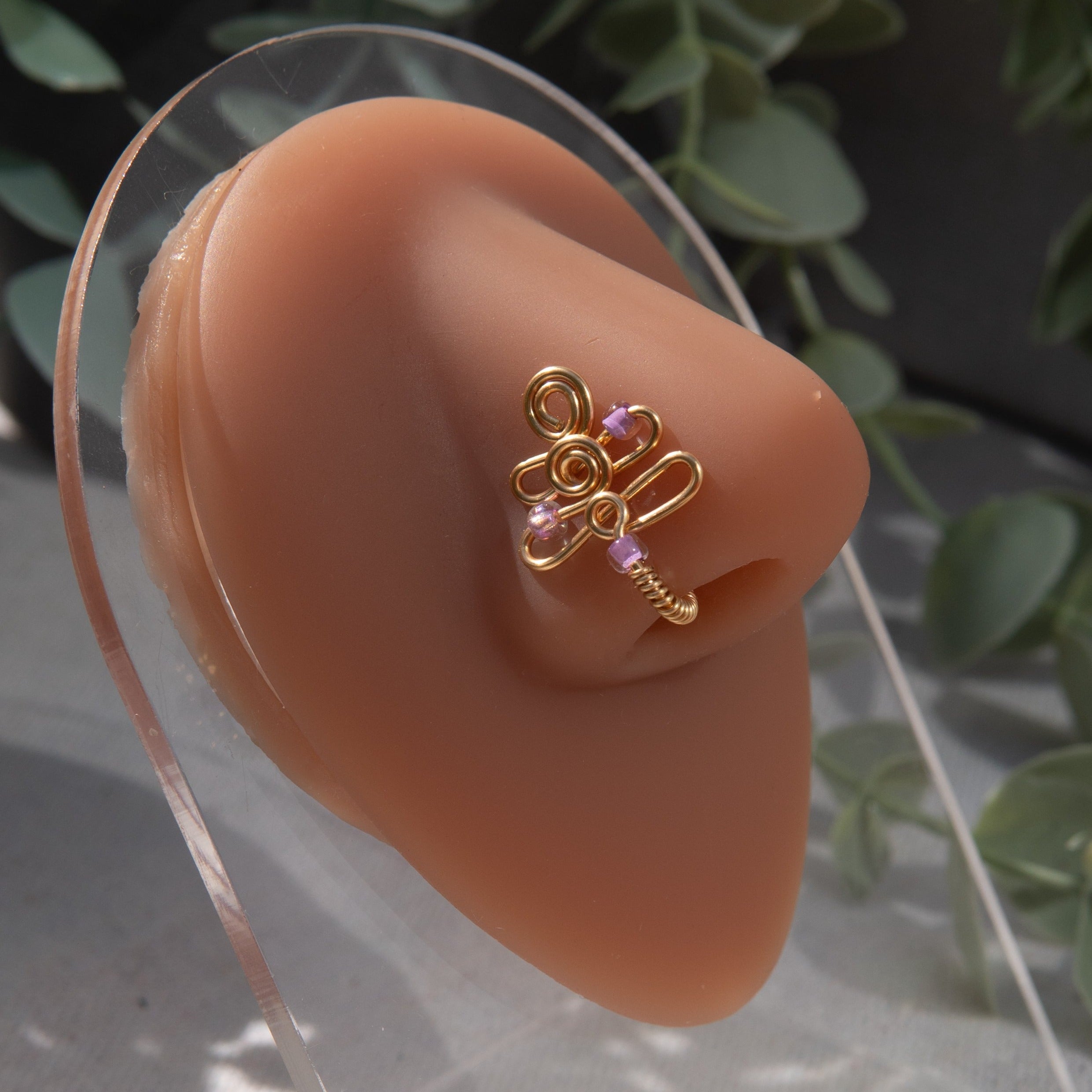 A gold-toned faux nose piercing cuffs the model's nostril. The wire design forms an abstract spiral shape with three lilac seed beads intertwined into the design.