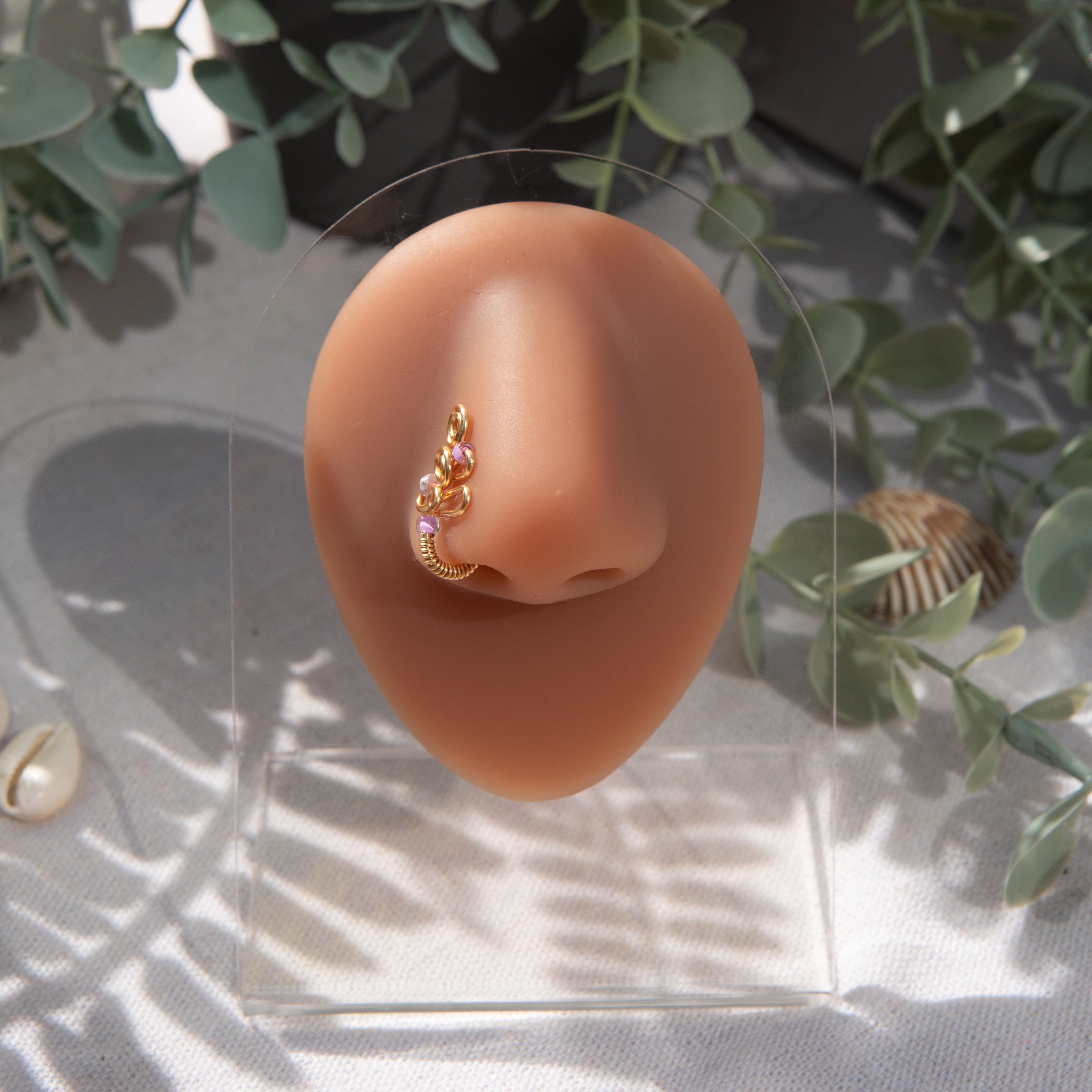 A gold-toned faux nose piercing cuffs the model's nostril. The wire design forms an abstract spiral shape with three lilac seed beads intertwined into the design.