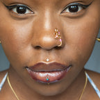 A gold-toned faux nose piercing cuffs the model's nostril. The wire design forms an abstract spiral shape with three pink seed beads intertwined into the design.