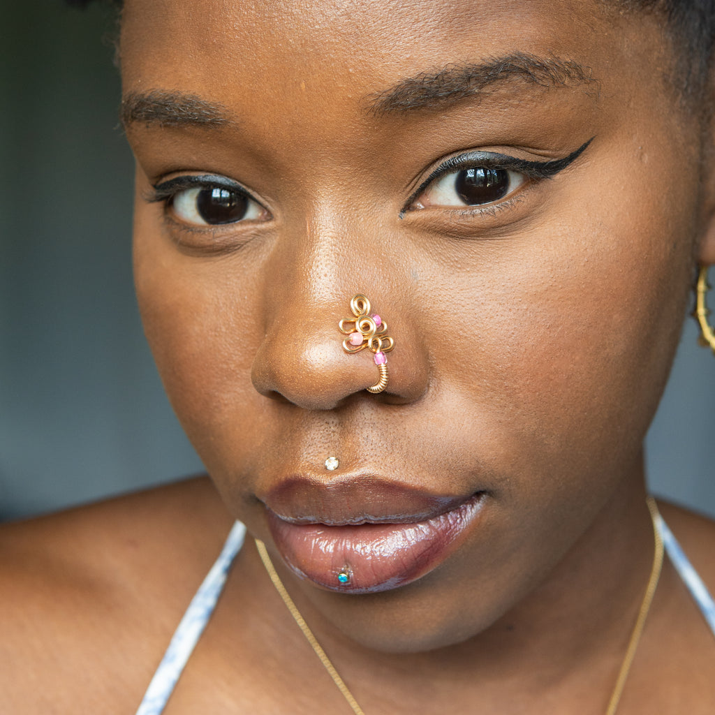 A gold-toned faux nose piercing cuffs the model's nostril. The wire design forms an abstract spiral shape with three pink seed beads intertwined into the design.