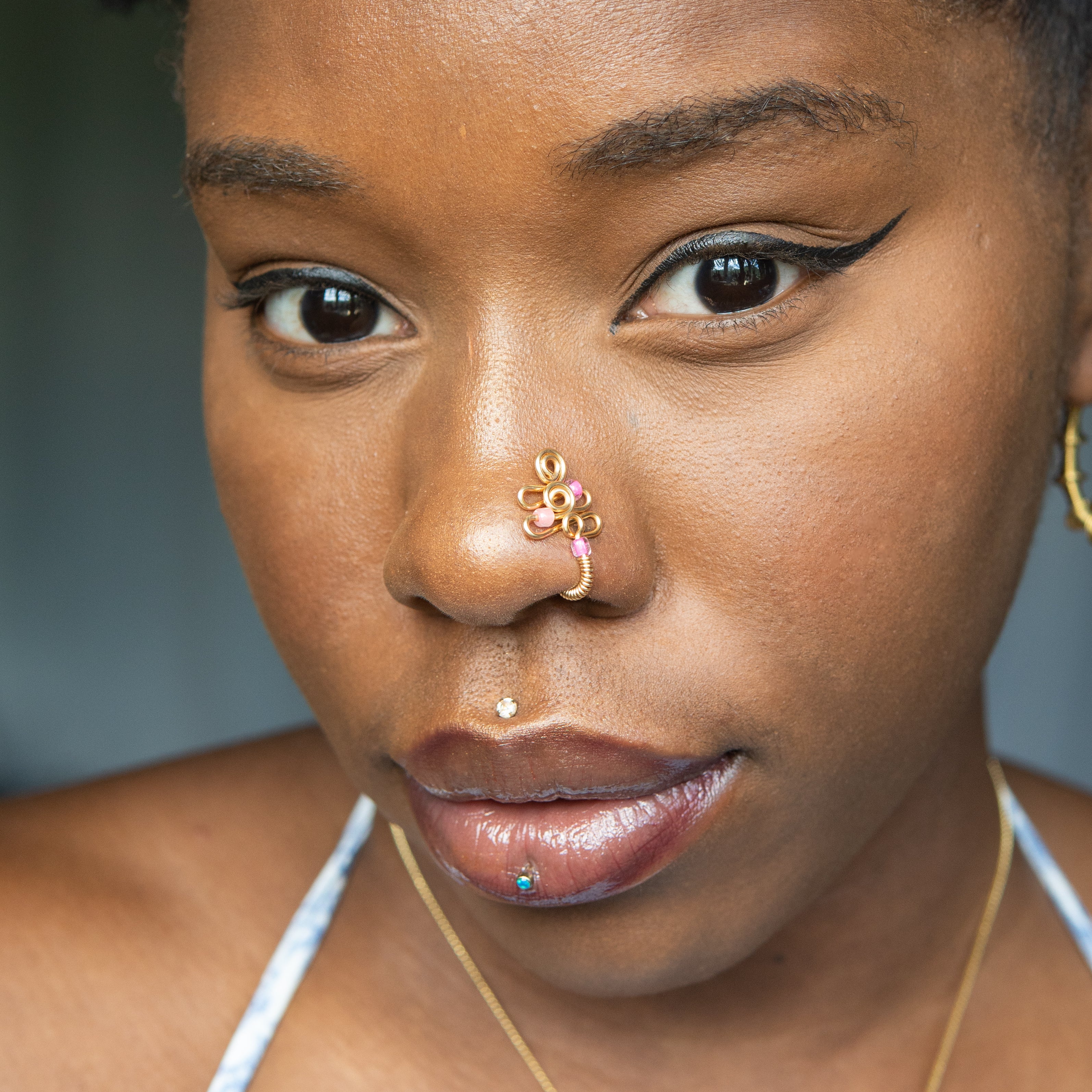 A gold-toned faux nose piercing cuffs the model's nostril. The wire design forms an abstract spiral shape with three pink seed beads intertwined into the design.