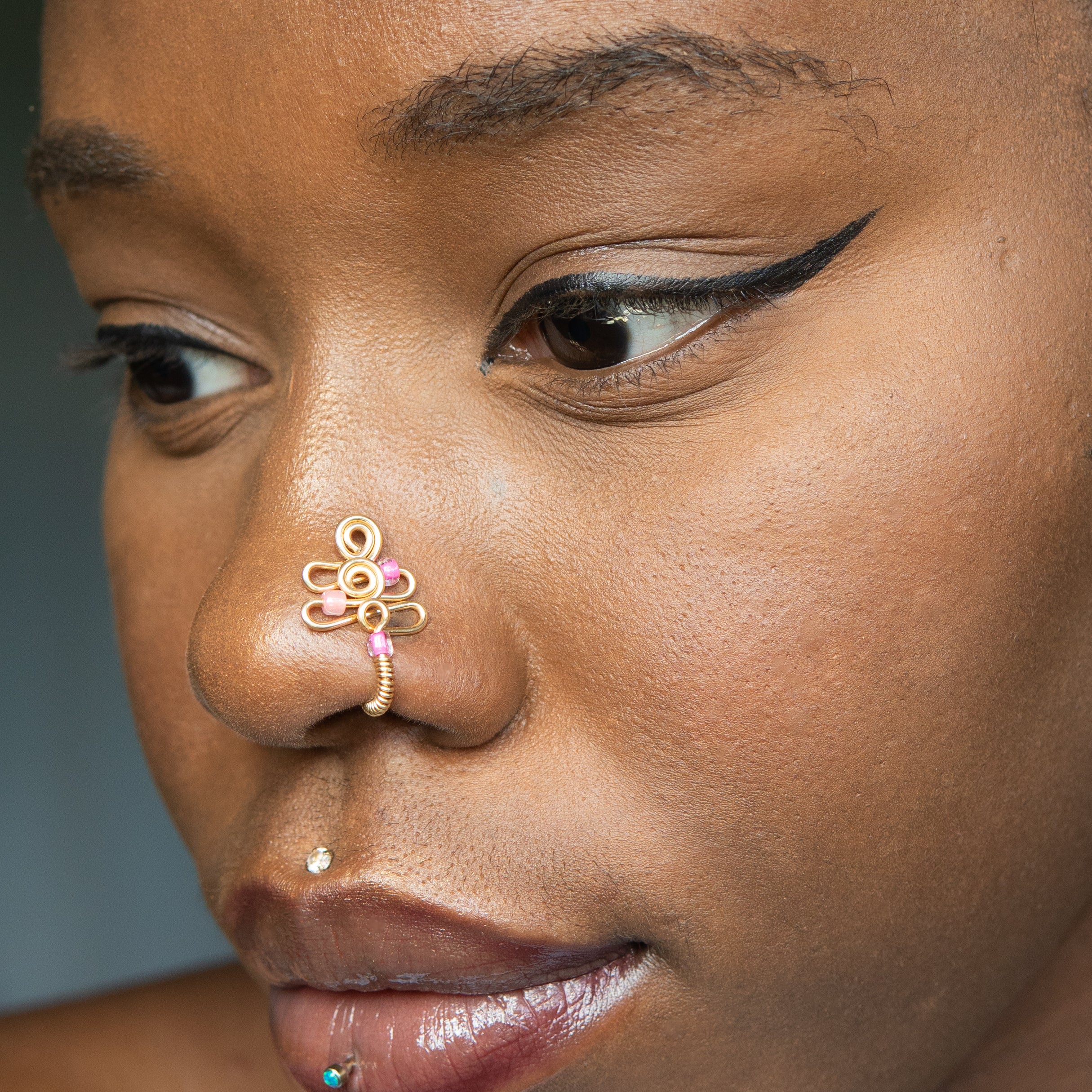 A gold-toned faux nose piercing cuffs the model's nostril. The wire design forms an abstract spiral shape with three pink seed beads intertwined into the design.