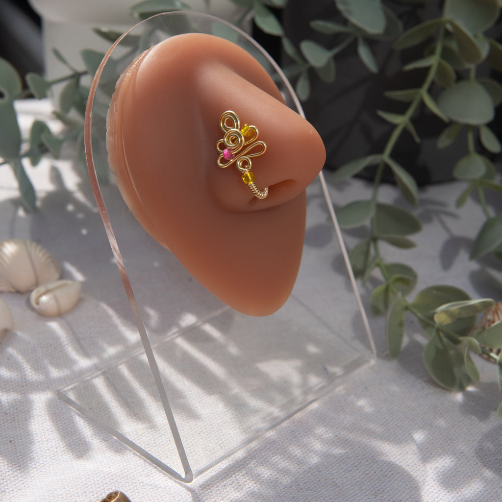 A light gold-toned faux nose piercing cuffs the model's nostril. The wire design forms an abstract spiral shape with three seed beads (yellow and pink) intertwined into the design.