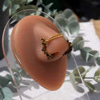 A bronze-toned chain bridge faux nose piercing cuffs both nostrils of the model. Brown transparent seed beads are wire-wrapped around the circumference of each cuff. Two dainty 18K gold-plated curb chains rest across the bridge of the nose attached to the top of each cuff.