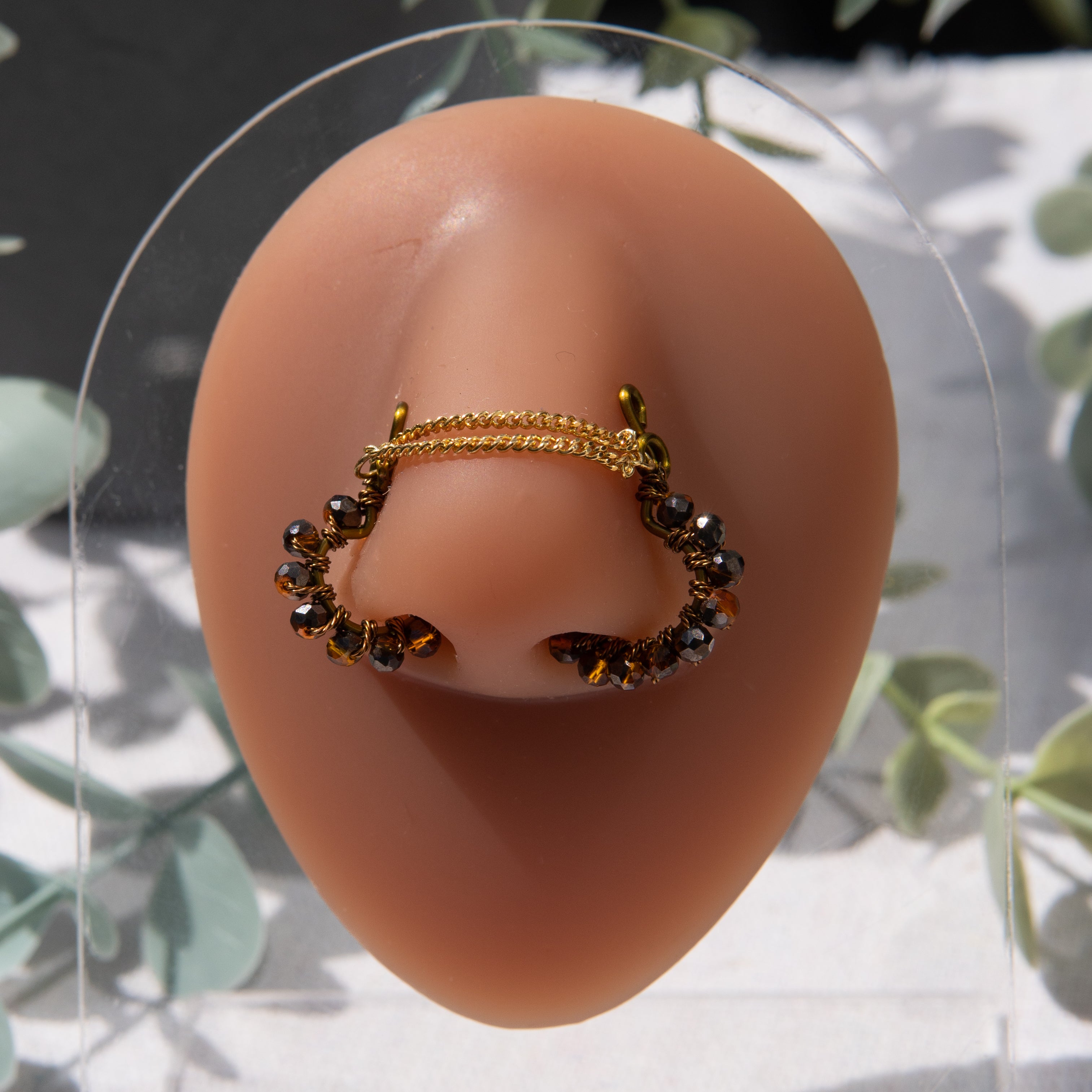 A bronze-toned chain bridge faux nose piercing cuffs both nostrils of the model. Brown transparent seed beads are wire-wrapped around the circumference of each cuff. Two dainty 18K gold-plated curb chains rest across the bridge of the nose attached to the top of each cuff.