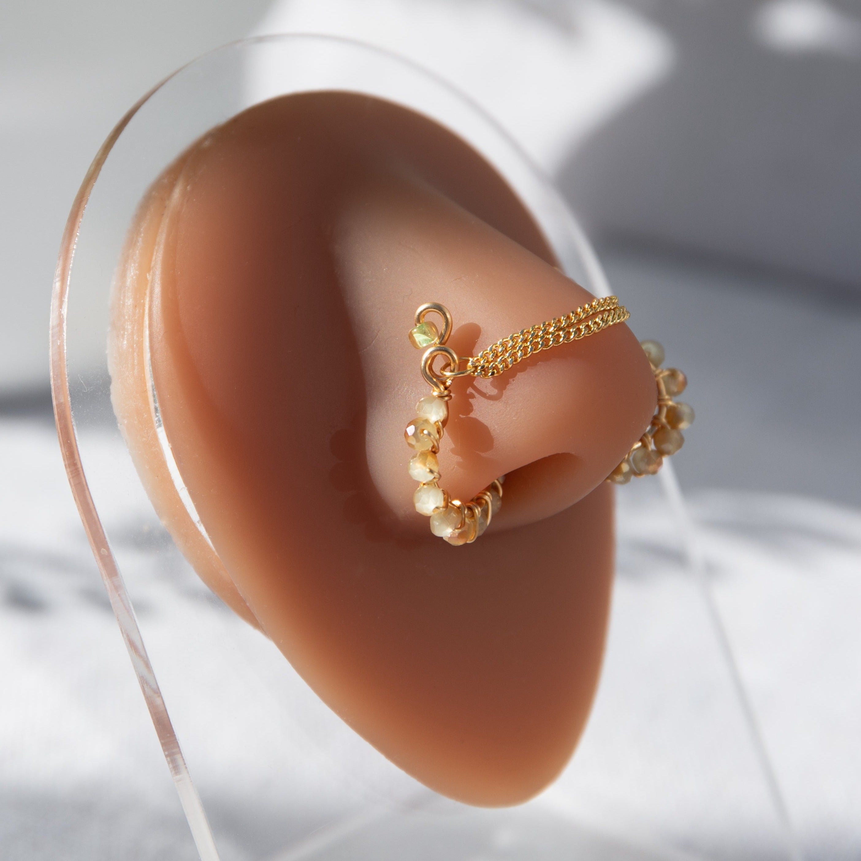 A gold-toned chain bridge faux nose ring cuffs both nostrils of the model. Beige AB seed beads are wire-wrapped around the circumference of each cuff. Two dainty 18K gold-plated curb chains rest across the bridge of the nose attached to the top of each cuff. 