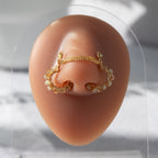A gold-toned chain bridge faux nose ring cuffs both nostrils of the model. Beige AB seed beads are wire-wrapped around the circumference of each cuff. Two dainty 18K gold-plated curb chains rest across the bridge of the nose attached to the top of each cuff.
