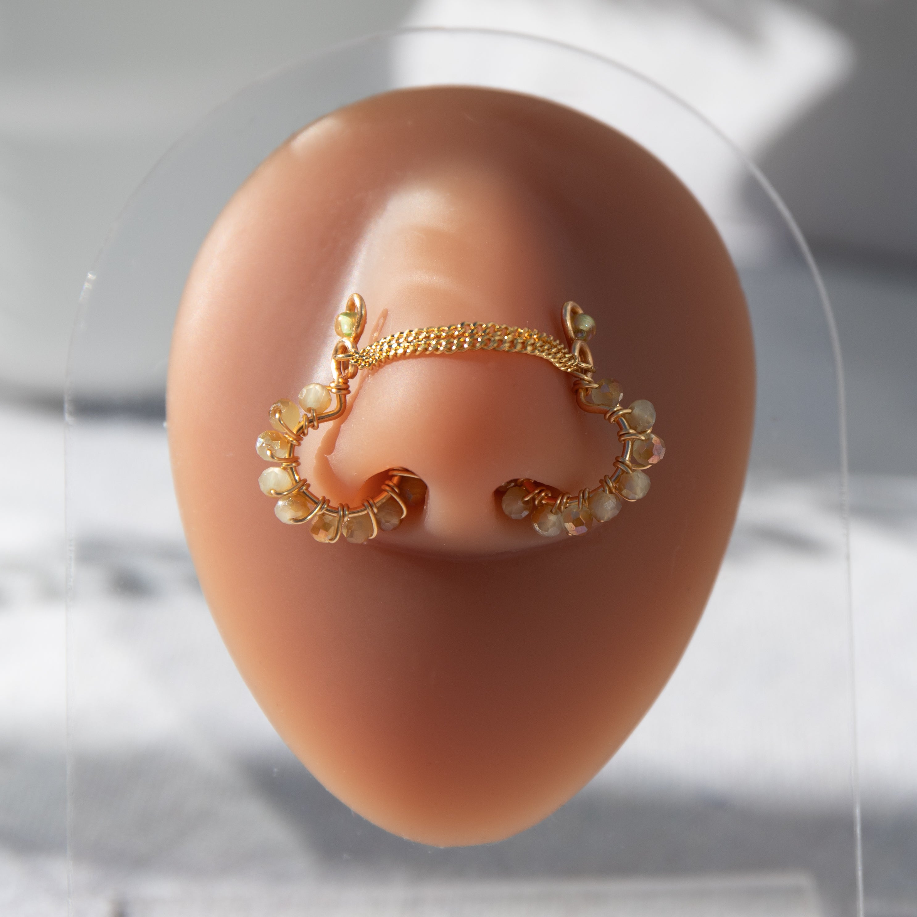 A gold-toned chain bridge faux nose ring cuffs both nostrils of the model. Beige AB seed beads are wire-wrapped around the circumference of each cuff. Two dainty 18K gold-plated curb chains rest across the bridge of the nose attached to the top of each cuff.