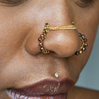 A bronze-toned chain bridge faux nose piercing cuffs both nostrils of the model. Brown transparent seed beads are wire-wrapped around the circumference of each cuff. Two dainty 18K gold-plated curb chains rest across the bridge of the nose attached to the top of each cuff.