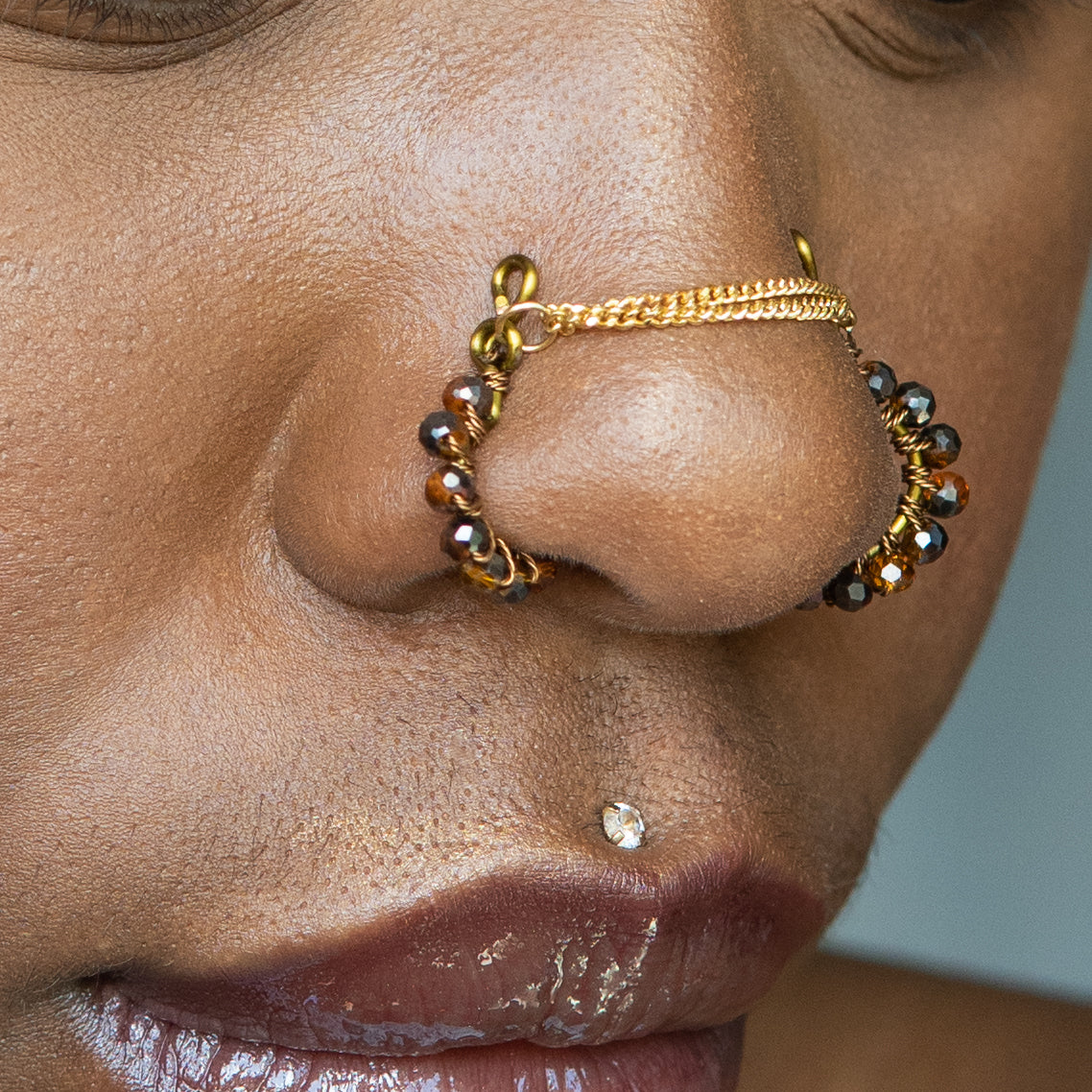 A bronze-toned chain bridge faux nose piercing cuffs both nostrils of the model. Brown transparent seed beads are wire-wrapped around the circumference of each cuff. Two dainty 18K gold-plated curb chains rest across the bridge of the nose attached to the top of each cuff.