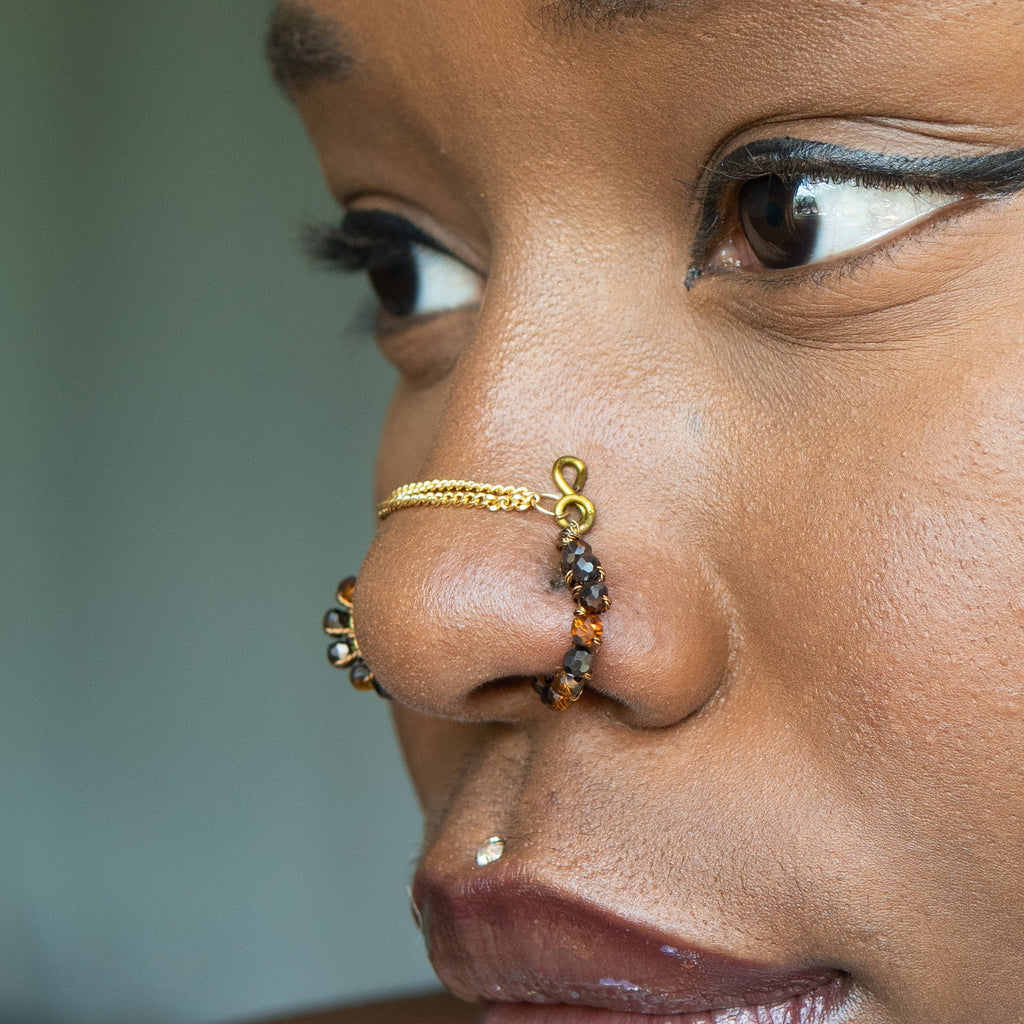 A bronze-toned chain bridge faux nose piercing cuffs both nostrils of the model. Brown transparent seed beads are wire-wrapped around the circumference of each cuff. Two dainty 18K gold-plated curb chains rest across the bridge of the nose attached to the top of each cuff.
