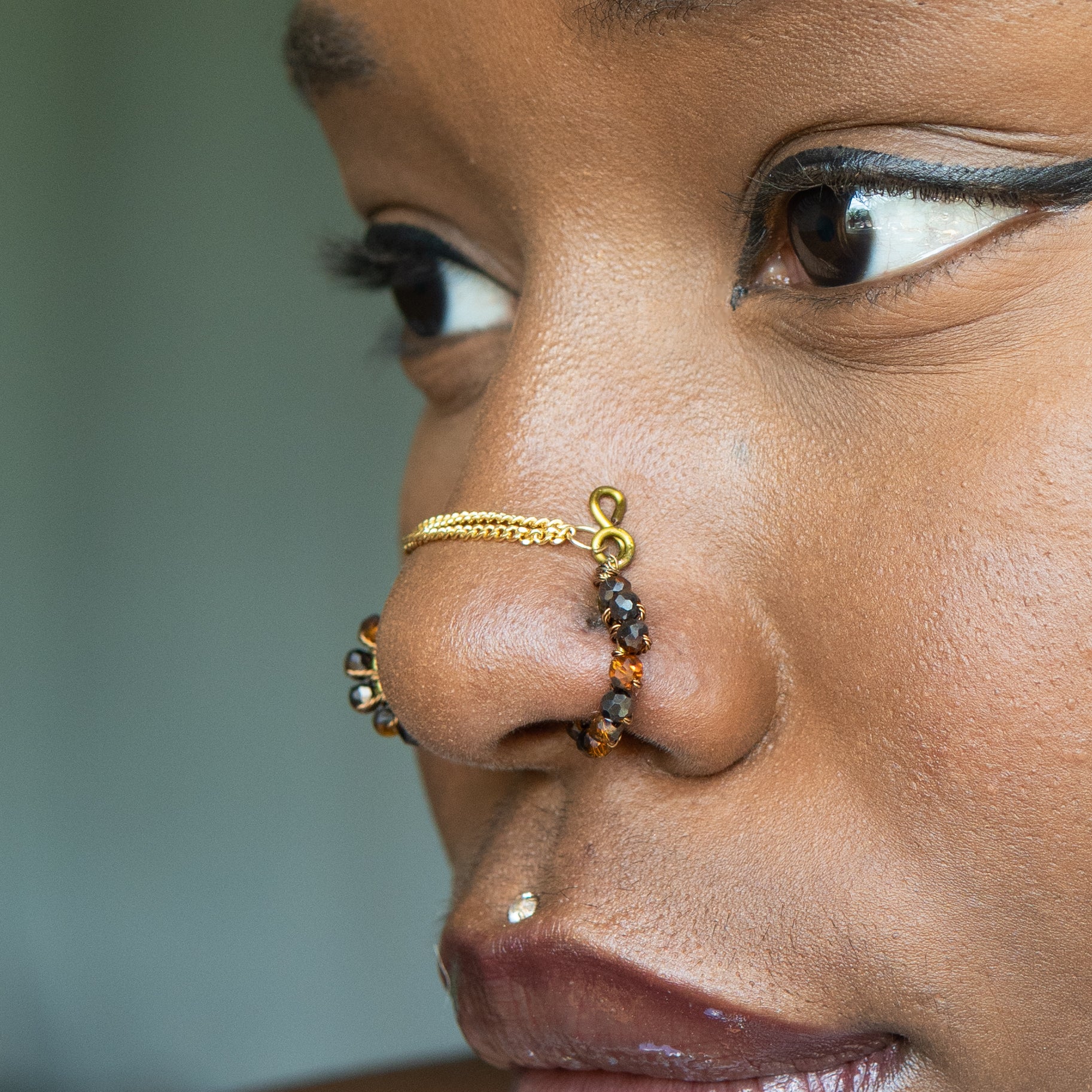 A bronze-toned chain bridge faux nose piercing cuffs both nostrils of the model. Brown transparent seed beads are wire-wrapped around the circumference of each cuff. Two dainty 18K gold-plated curb chains rest across the bridge of the nose attached to the top of each cuff.