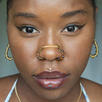 A bronze-toned chain bridge faux nose piercing cuffs both nostrils of the model. Brown transparent seed beads are wire-wrapped around the circumference of each cuff. Two dainty 18K gold-plated curb chains rest across the bridge of the nose attached to the top of each cuff.