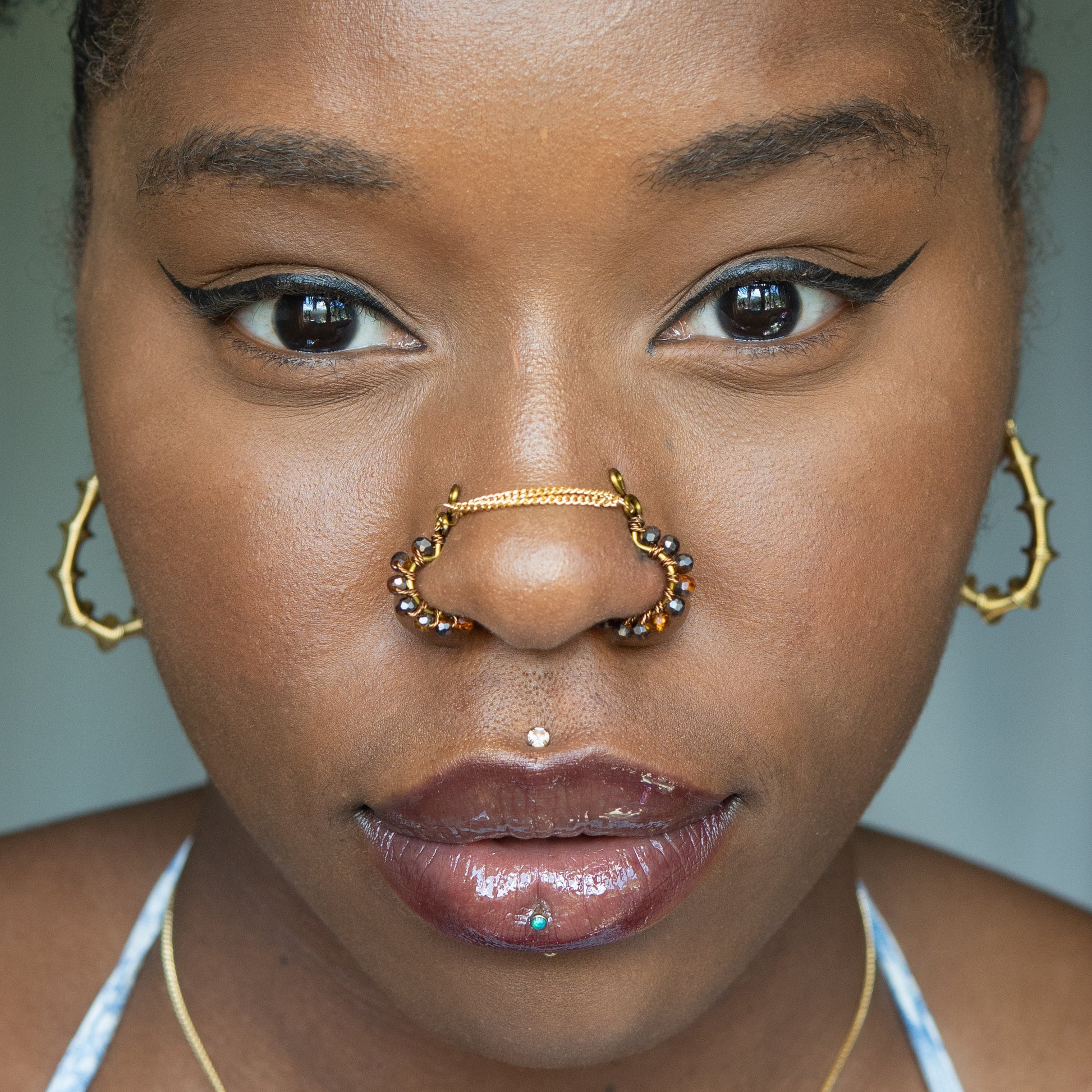 A bronze-toned chain bridge faux nose piercing cuffs both nostrils of the model. Brown transparent seed beads are wire-wrapped around the circumference of each cuff. Two dainty 18K gold-plated curb chains rest across the bridge of the nose attached to the top of each cuff.