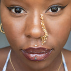 A gold-toned faux dangle nose ring cuffs the model's nostrils. Ombre nude seed beads are wire-wrapped around the hoop's circumference, with three beaded gold chains dangling down. An elaborate wire design tightly spirals up the nose, decreasing in size, with two additional dangle chains.