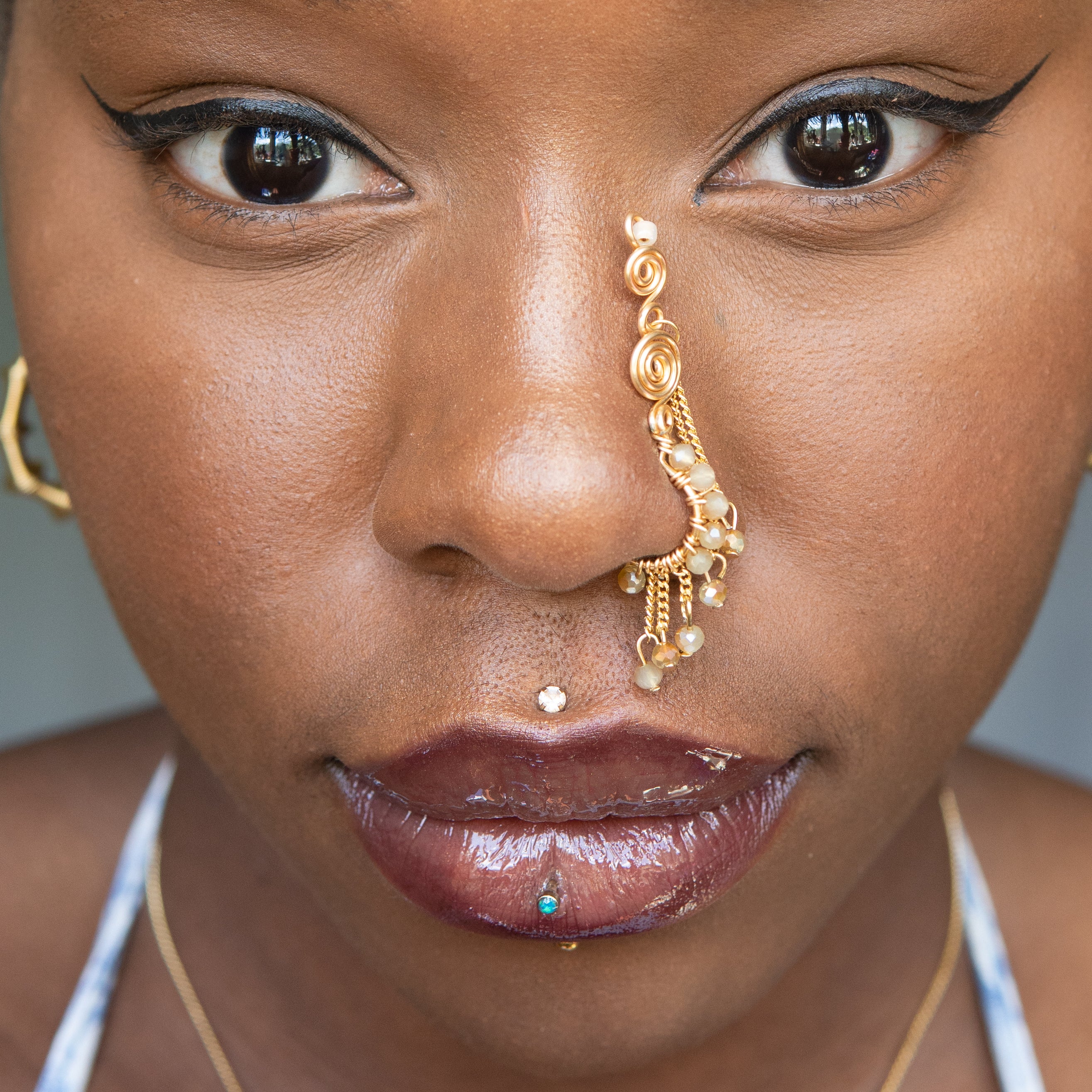 A gold-toned faux dangle nose ring cuffs the model's nostrils. Ombre nude seed beads are wire-wrapped around the hoop's circumference, with three beaded gold chains dangling down. An elaborate wire design tightly spirals up the nose, decreasing in size, with two additional dangle chains.