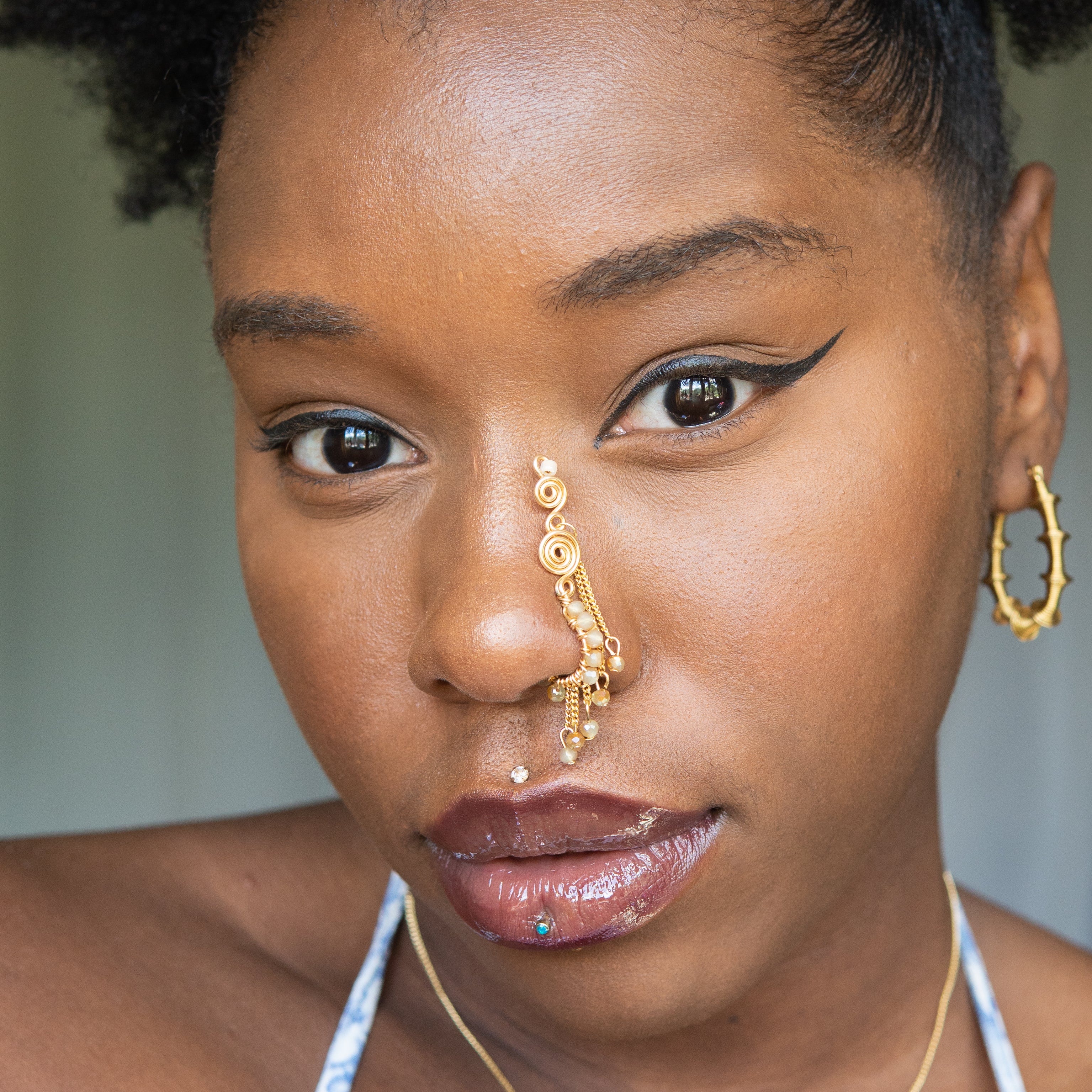 A gold-toned faux dangle nose ring cuffs the model's nostrils. Ombre nude seed beads are wire-wrapped around the hoop's circumference, with three beaded gold chains dangling down. An elaborate wire design tightly spirals up the nose, decreasing in size, with two additional dangle chains.