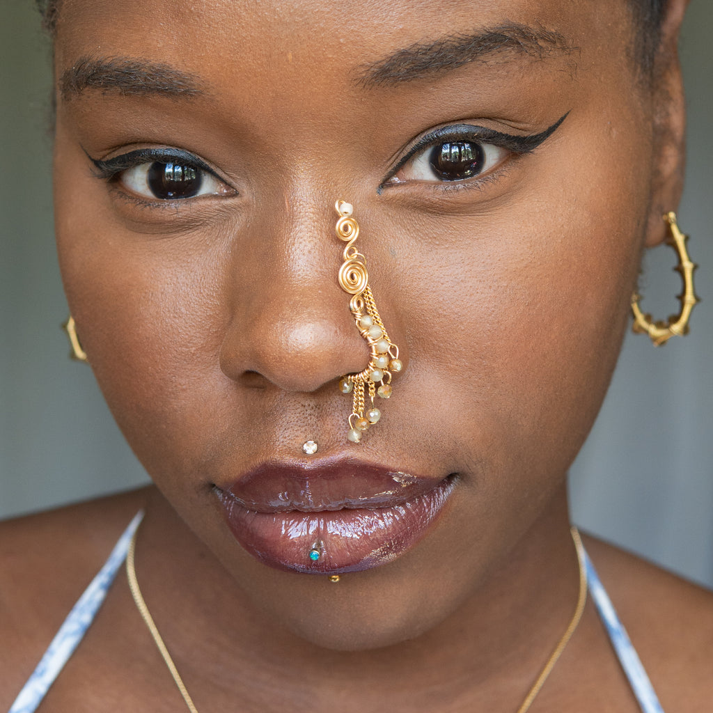 A gold-toned faux dangle nose ring cuffs the model's nostrils. Ombre nude seed beads are wire-wrapped around the hoop's circumference, with three beaded gold chains dangling down. An elaborate wire design tightly spirals up the nose, decreasing in size, with two additional dangle chains.