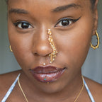 A gold-toned faux dangle nose ring cuffs the model's nostrils. Ombre nude seed beads are wire-wrapped around the hoop's circumference, with three beaded gold chains dangling down. An elaborate wire design tightly spirals up the nose, decreasing in size, with two additional dangle chains.