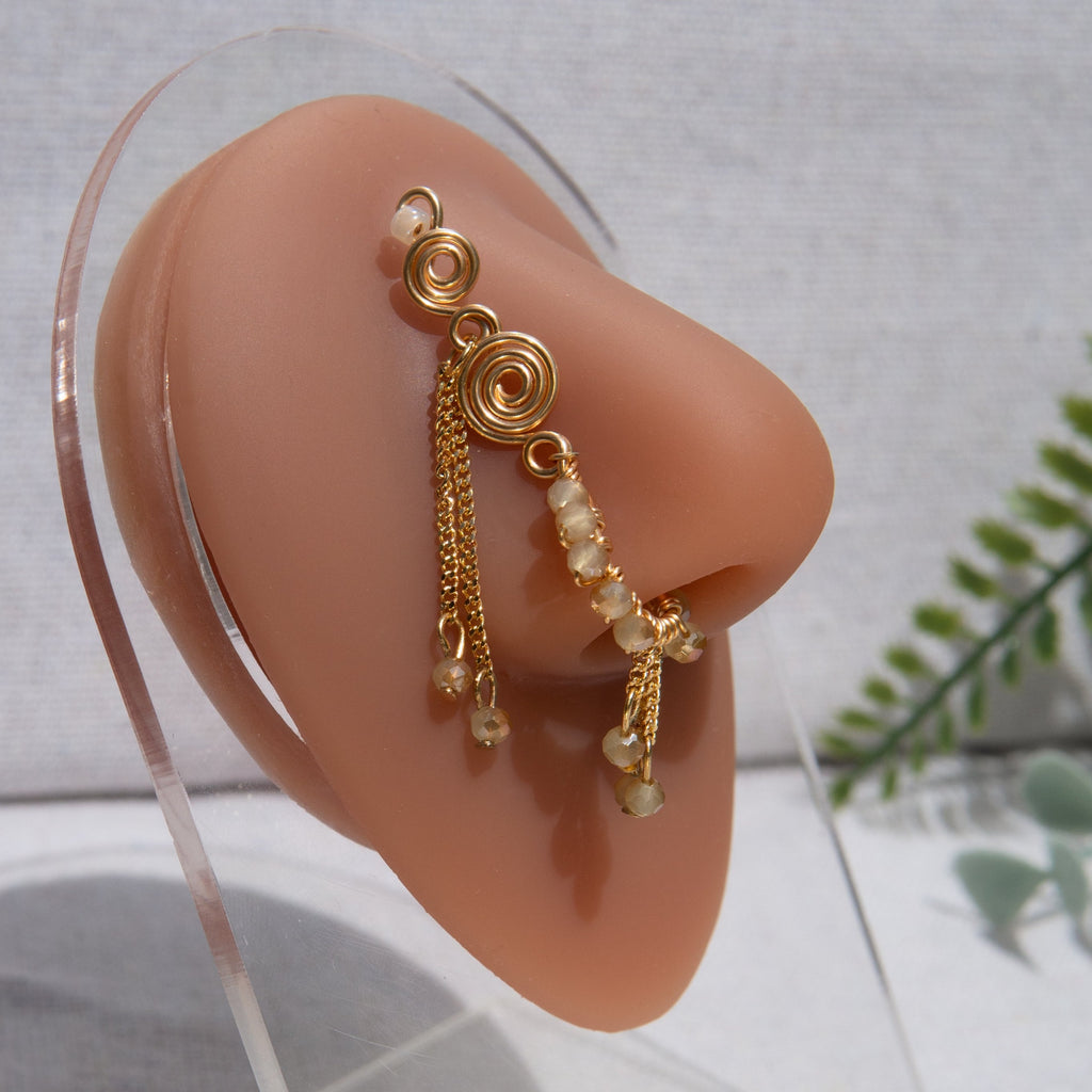 A gold-toned faux dangle nose ring cuffs the model's nostrils. Ombre nude seed beads are wire-wrapped around the hoop's circumference, with three beaded gold chains dangling down. An elaborate wire design tightly spirals up the nose, decreasing in size, with two additional dangle chains. 