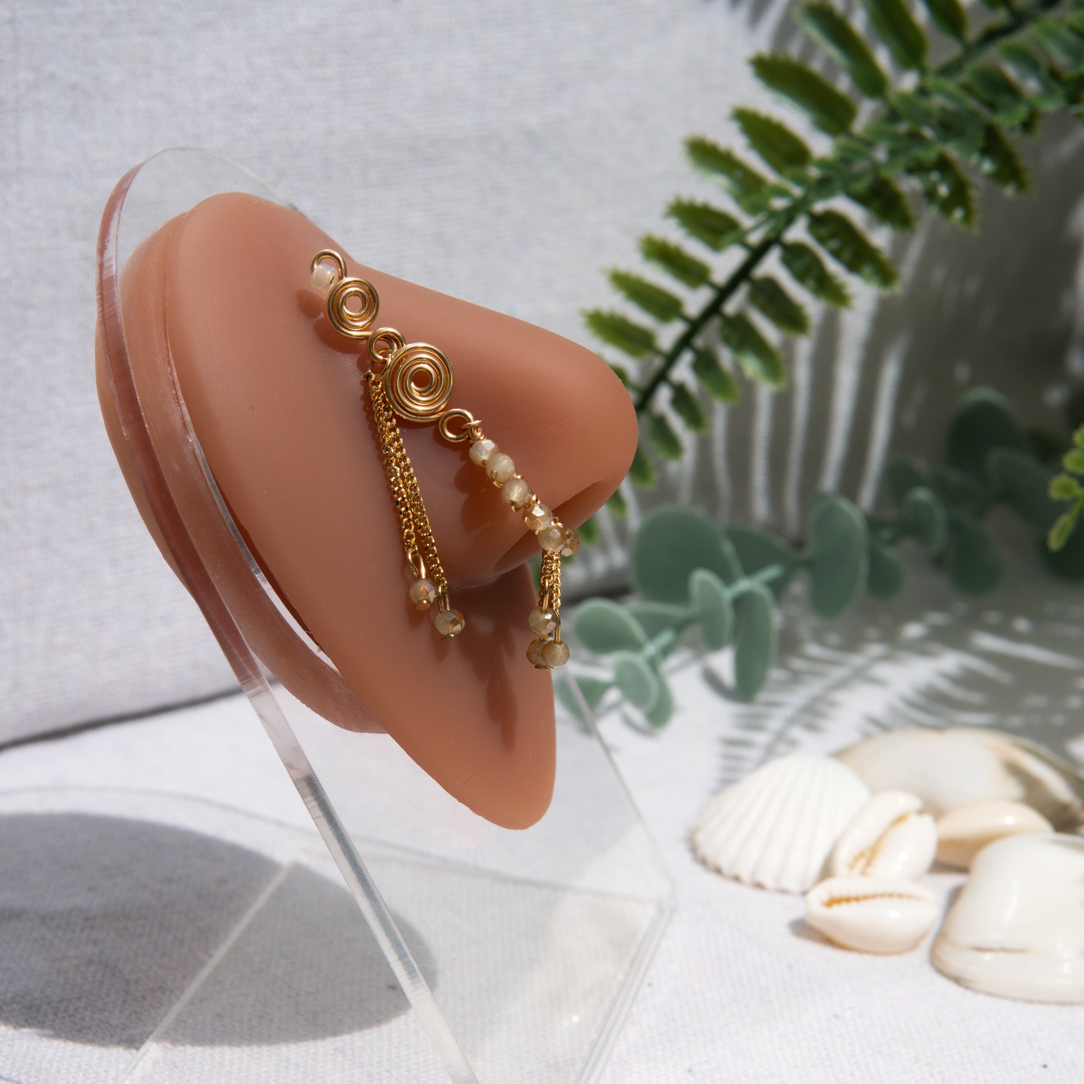 A gold-toned faux dangle nose ring cuffs the model's nostrils. Ombre nude seed beads are wire-wrapped around the hoop's circumference, with three beaded gold chains dangling down. An elaborate wire design tightly spirals up the nose, decreasing in size, with two additional dangle chains.