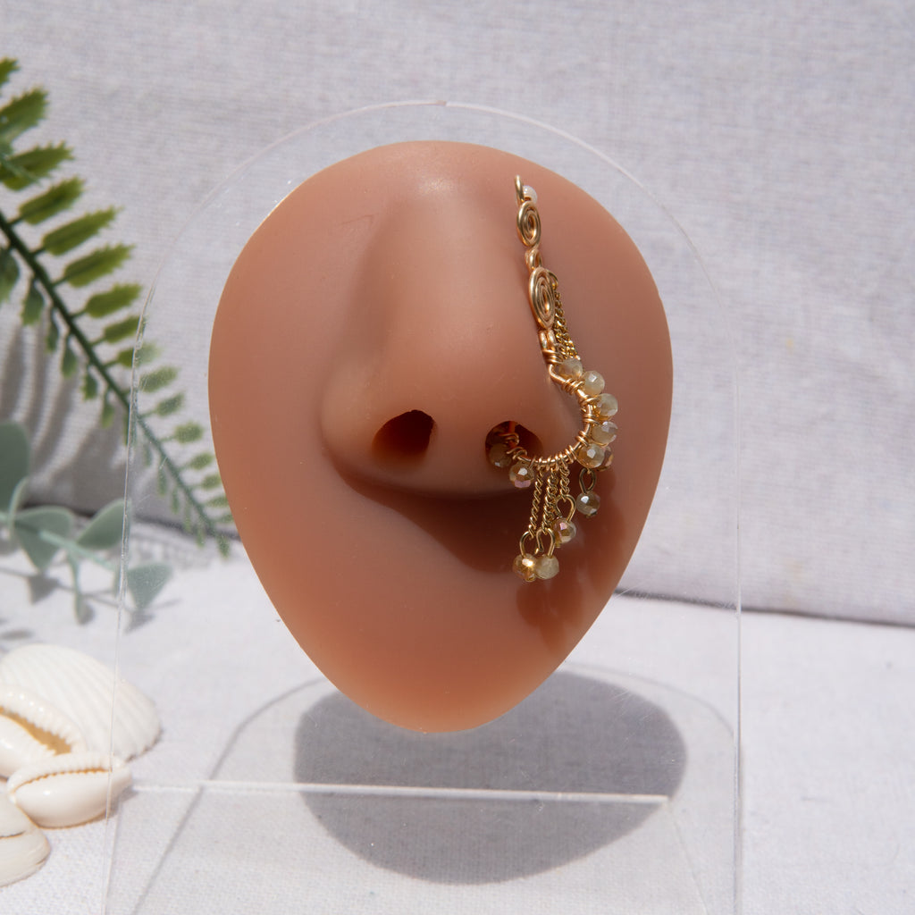 A gold-toned faux dangle nose ring cuffs the model's nostrils. Ombre nude seed beads are wire-wrapped around the hoop's circumference, with three beaded gold chains dangling down. An elaborate wire design tightly spirals up the nose, decreasing in size, with two additional dangle chains.