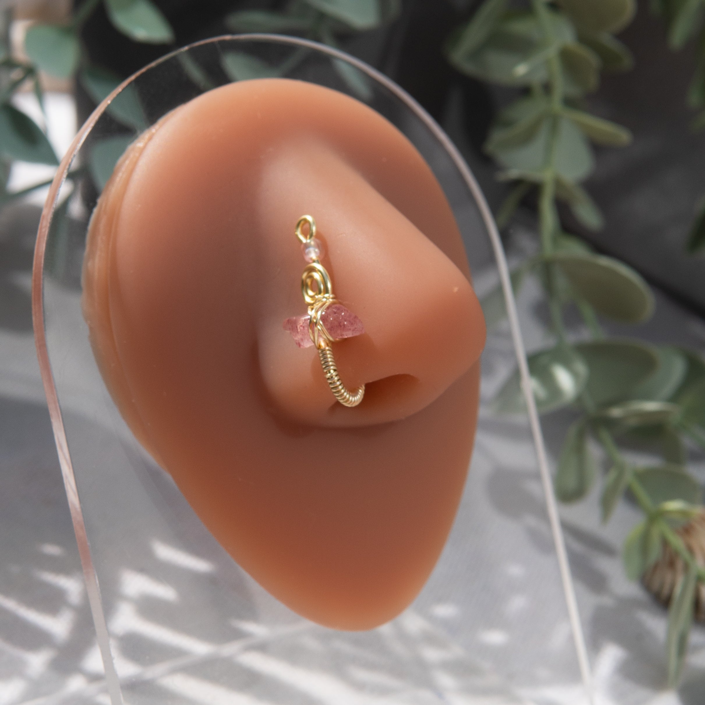 A light gold-toned faux nose ring cuffs the model's nostril. A pink sunstone crystal gemstone is wire-wrapped and interweaved into a vertical spiral wire design. A transparent pink seed bead resides at the top of the cuff design. 