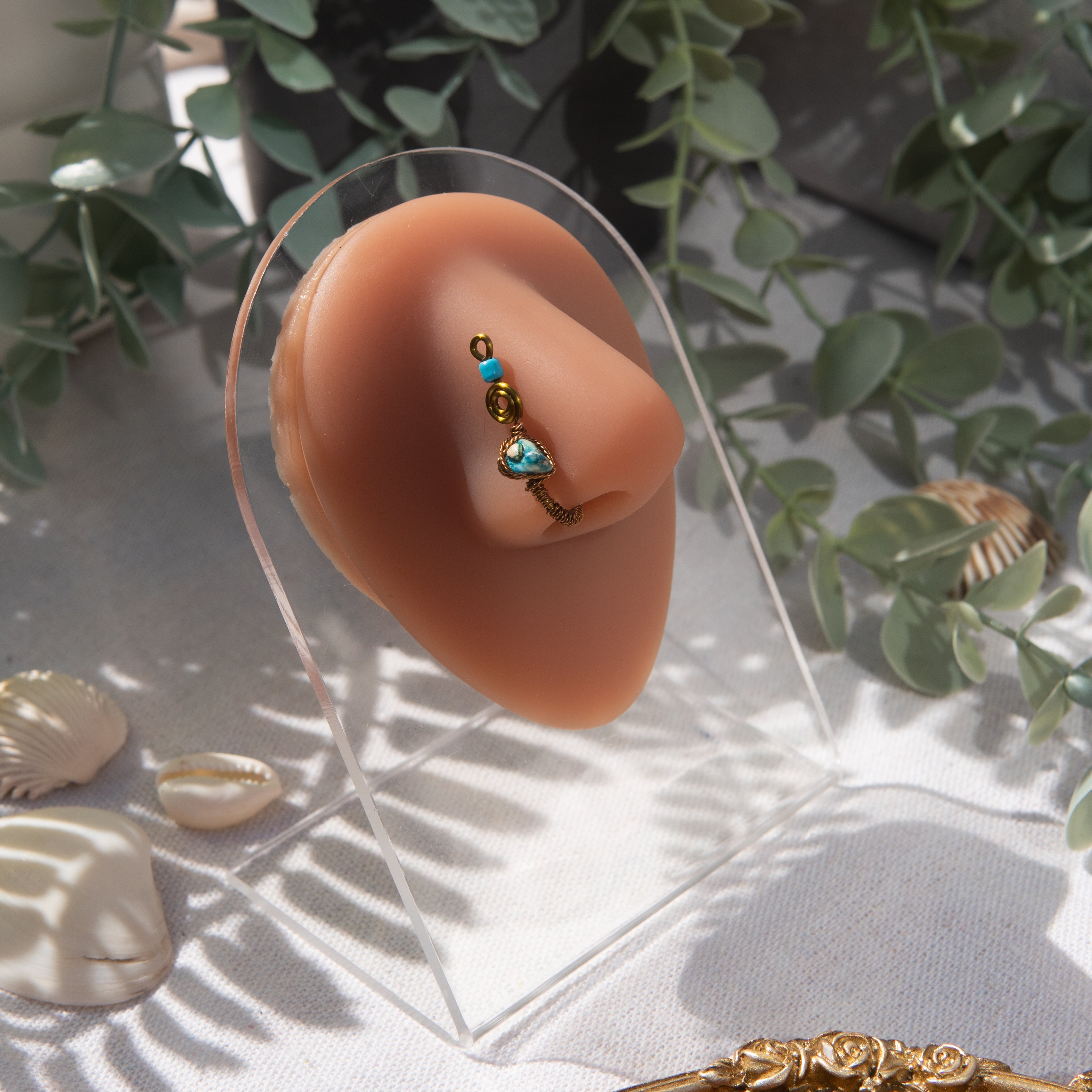 A bronze-toned faux nose ring cuffs the model's nostril. A turquoise crystal gemstone is wire-wrapped and interweaved into a vertical spiral wire design. A turquoise seed bead resides at the top of the cuff design. 