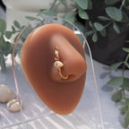 A gold-toned faux nose ring cuffs the model's nostril. A white shell chip is wire-wrapped and interweaved into a vertical spiral wire design. A transparent beige seed bead resides at the top of the cuff design.