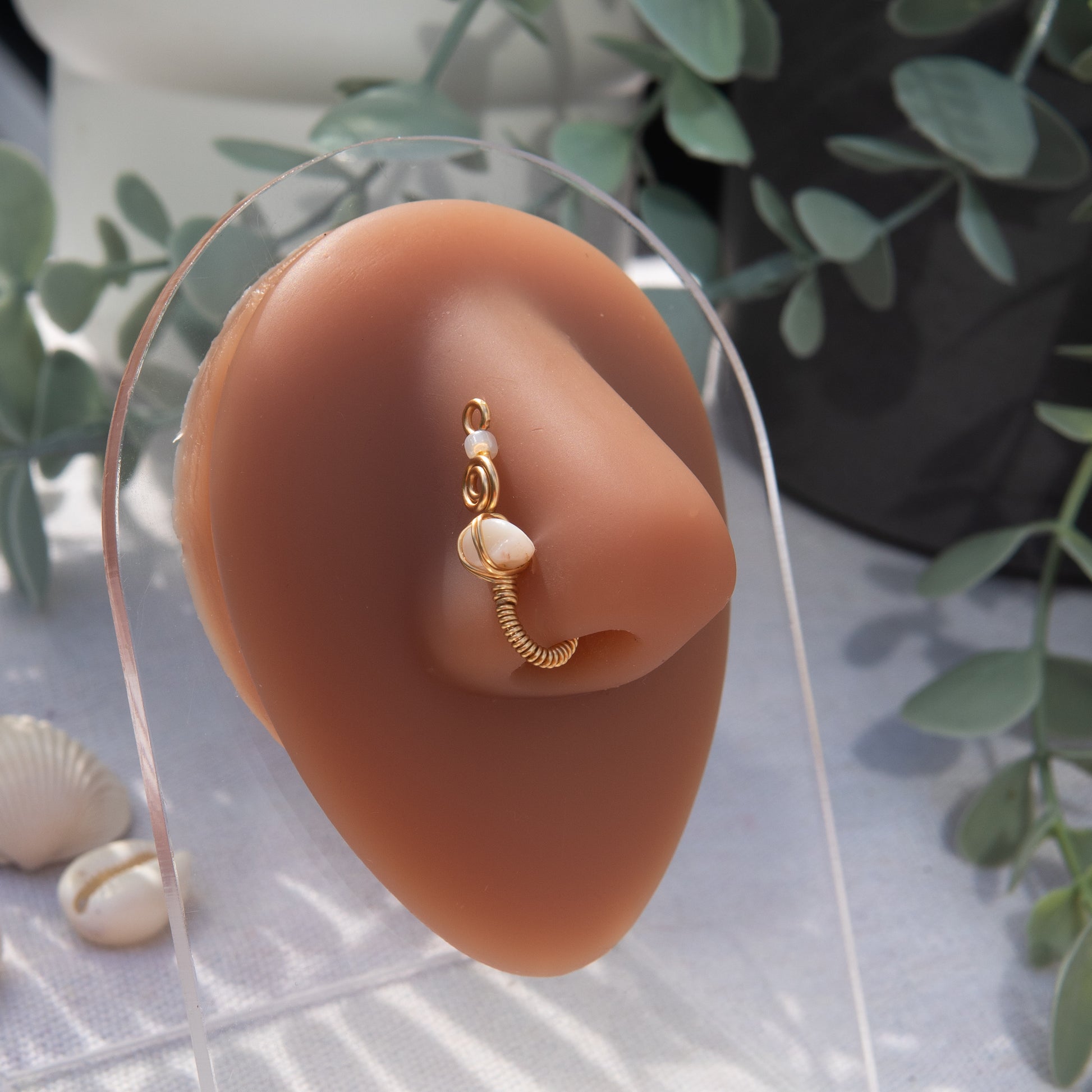 A gold-toned faux nose ring cuffs the model's nostril. A white shell chip is wire-wrapped and interweaved into a vertical spiral wire design. A transparent beige seed bead resides at the top of the cuff design.