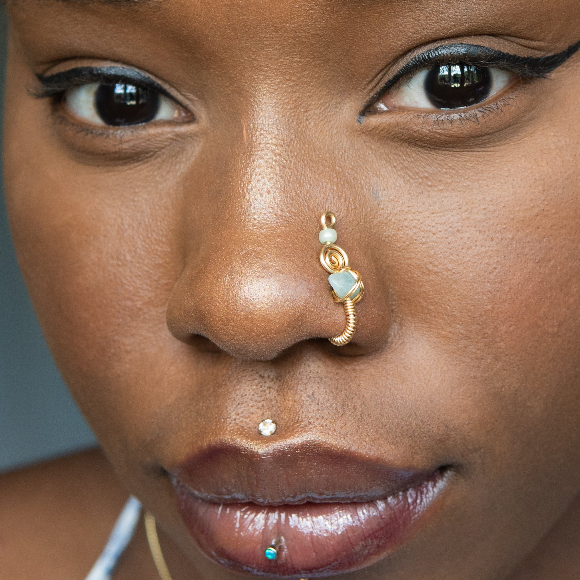  A gold-toned faux nose ring cuffs the model's nostril. A green aventurine crystal gemstone is wire-wrapped and interweaved into a vertical spiral wire design.
