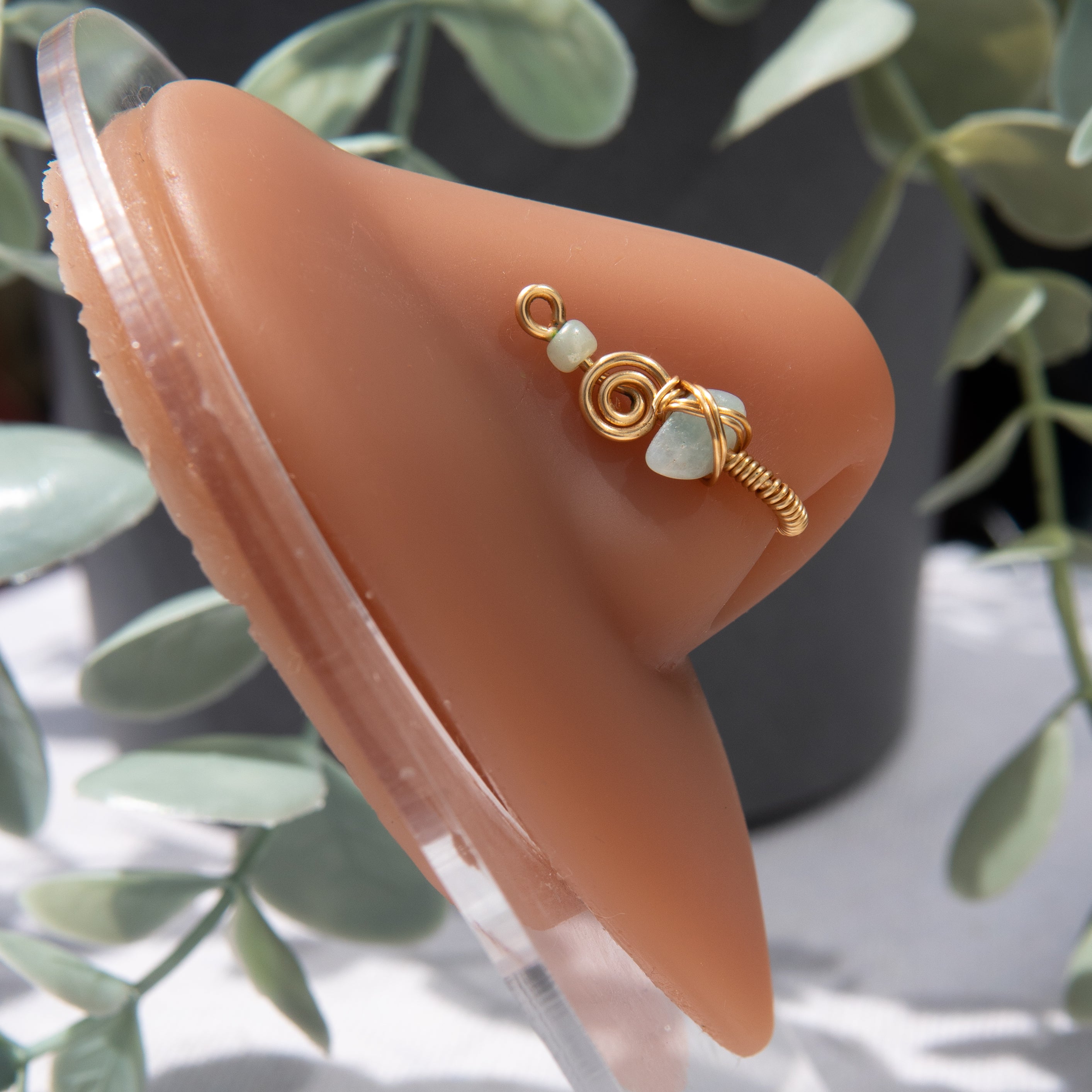 A gold-toned faux nose ring cuffs the model's nostril. A green aventurine crystal gemstone is wire-wrapped and interweaved into a vertical spiral wire design. A green seed bead resides at the top of the cuff design. 
