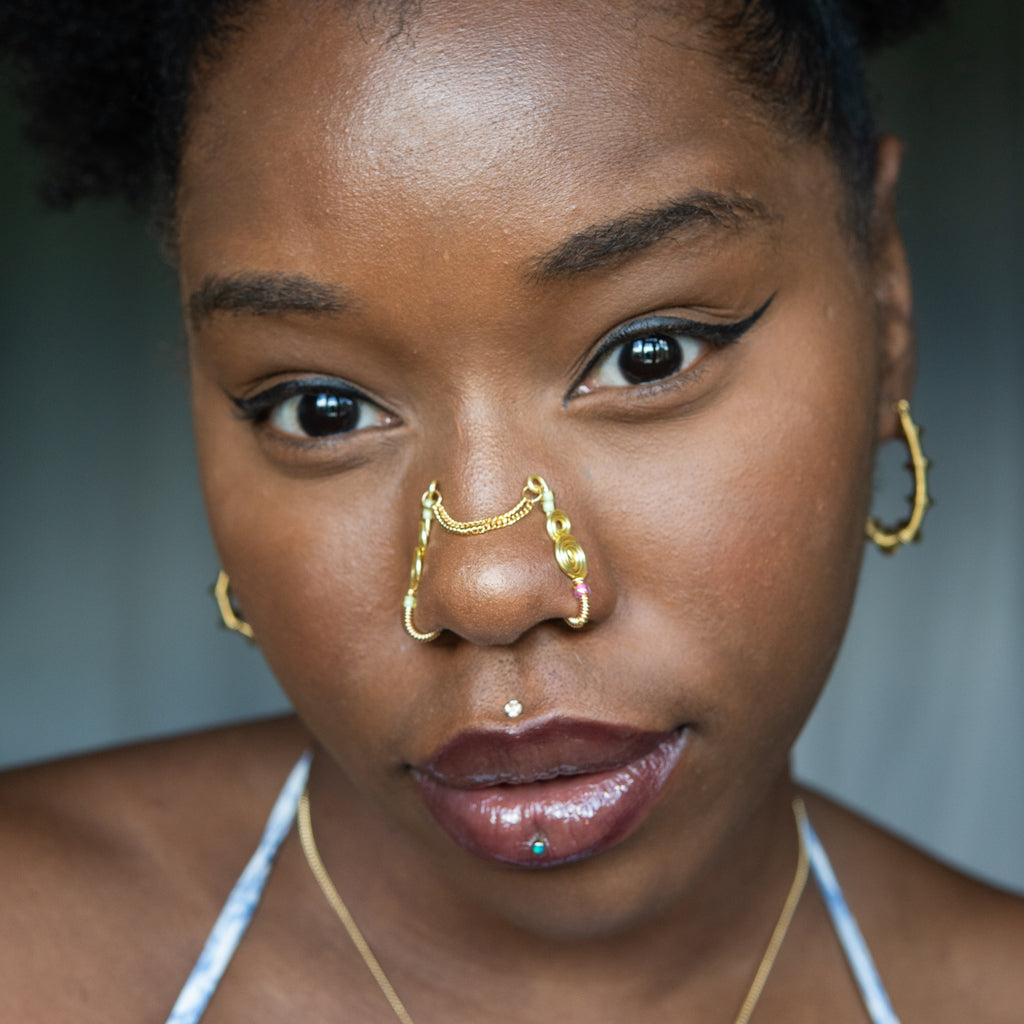 A two-toned faux bridge nose piercing in gold cuffs both nostrils of the model. Two dainty 18K gold-plated curb chains rest across the bridge of the nose, attached to the top of each nose cuff. Three seed beads are interweaved into the design of each nose cuff.