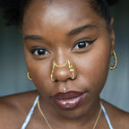 A two-toned faux bridge nose piercing in gold cuffs both nostrils of the model. Two dainty 18K gold-plated curb chains rest across the bridge of the nose, attached to the top of each nose cuff. Three seed beads are interweaved into the design of each nose cuff.