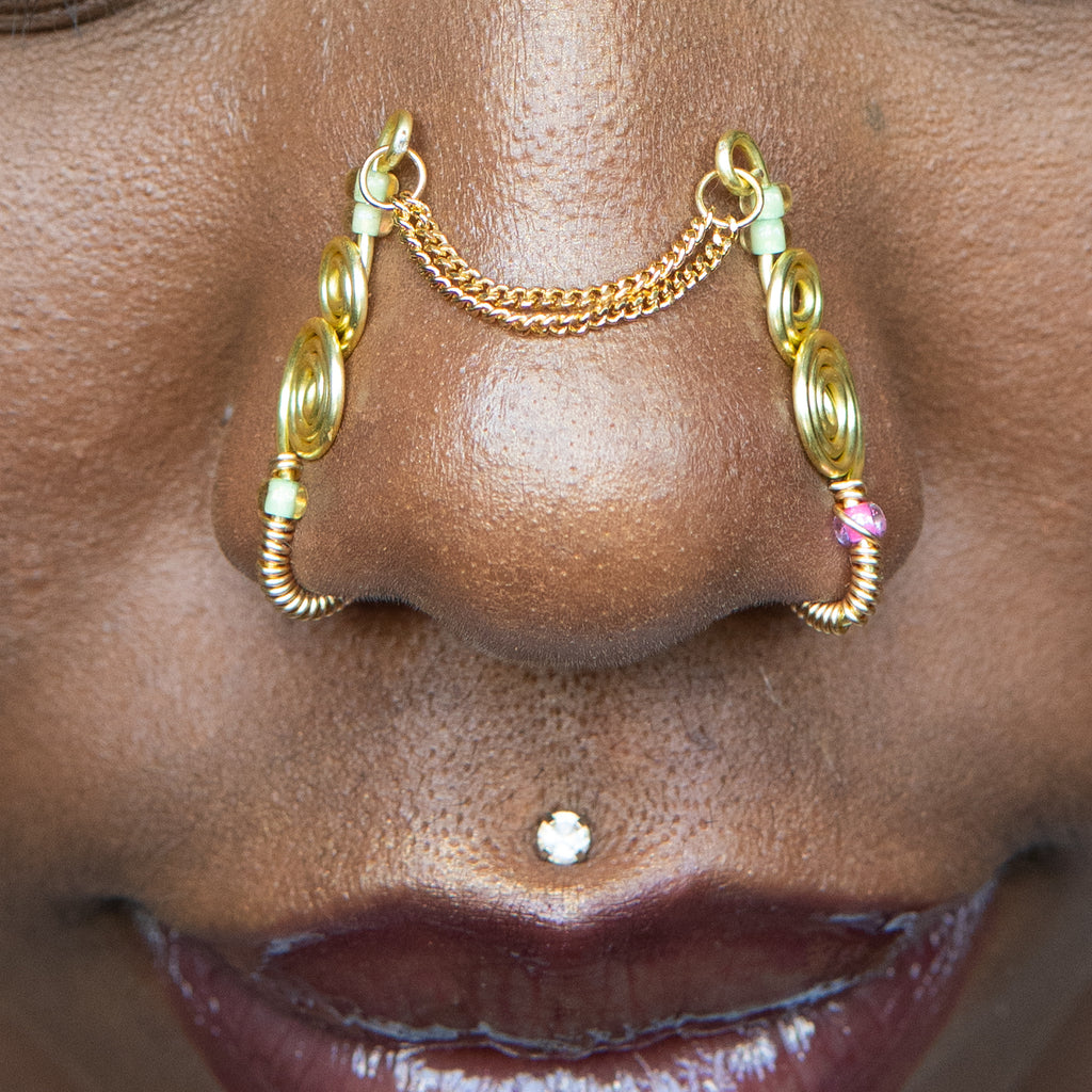 A two-toned faux bridge nose piercing in gold cuffs both nostrils of the model. Two dainty 18K gold-plated curb chains rest across the bridge of the nose, attached to the top of each nose cuff. Three seed beads are interweaved into the design of each nose cuff.