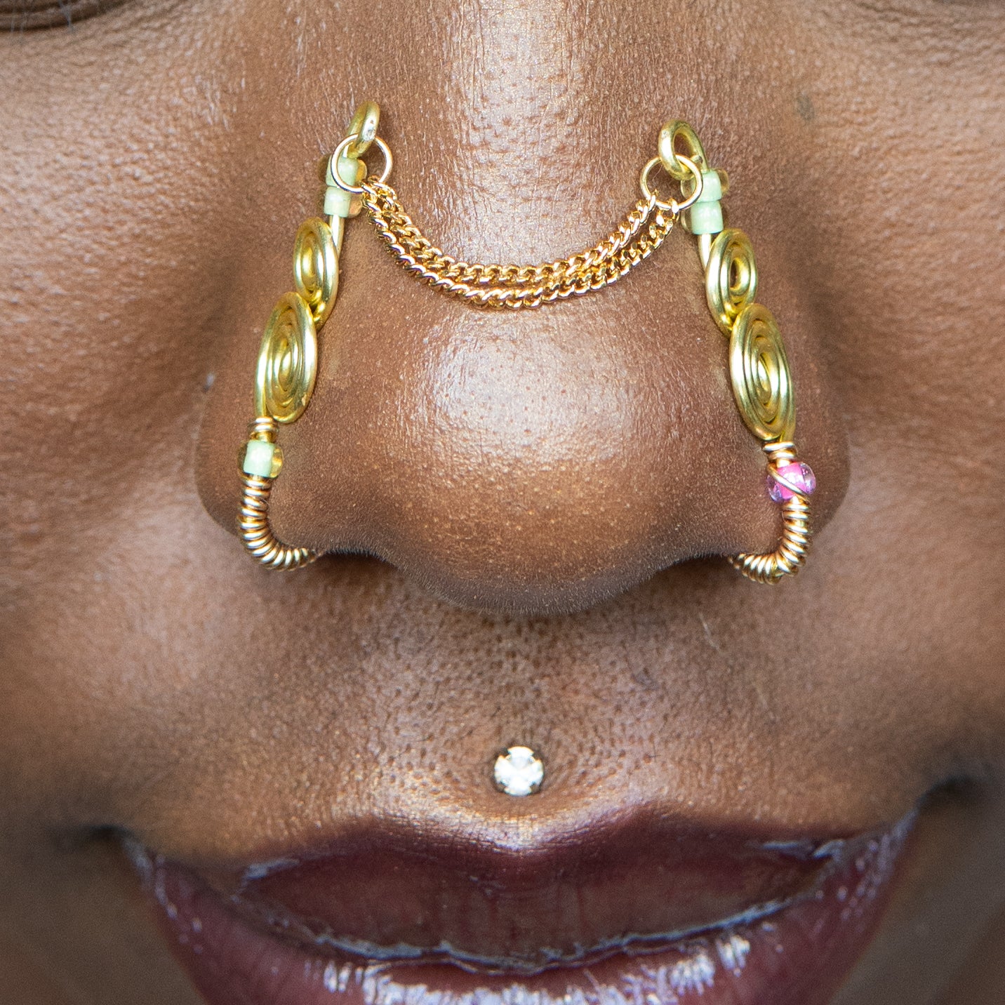 A two-toned faux bridge nose piercing in gold cuffs both nostrils of the model. Two dainty 18K gold-plated curb chains rest across the bridge of the nose, attached to the top of each nose cuff. Three seed beads are interweaved into the design of each nose cuff.