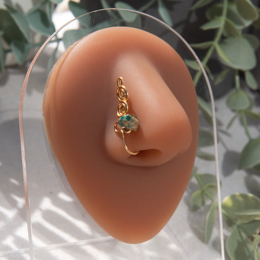 A gold-toned faux nose ring adorns the model's nose in a circular wire design that spirals up and gradually decreases in size. A crystal amazonite gemstone is woven into the design and rests at the bottom.