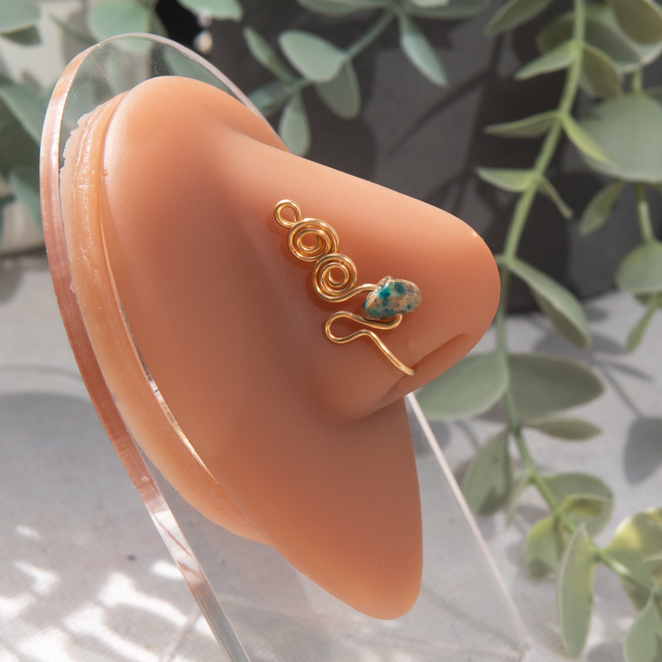 A gold-toned faux nose ring adorns the model's nose in a circular wire design that spirals up and gradually decreases in size. A crystal amazonite gemstone is woven into the design and rests at the bottom.