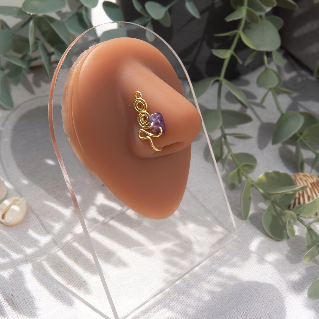 A gold-toned faux nose ring adorns the model's nose in a circular wire design that spirals up and gradually decreases in size. A crystal amethyst gemstone is woven into the design and rests at the bottom.