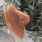 A gold-toned faux nose ring adorns the model's nose in a circular wire design that spirals up and gradually decreases in size. A crystal amethyst gemstone is woven into the design and rests at the bottom.