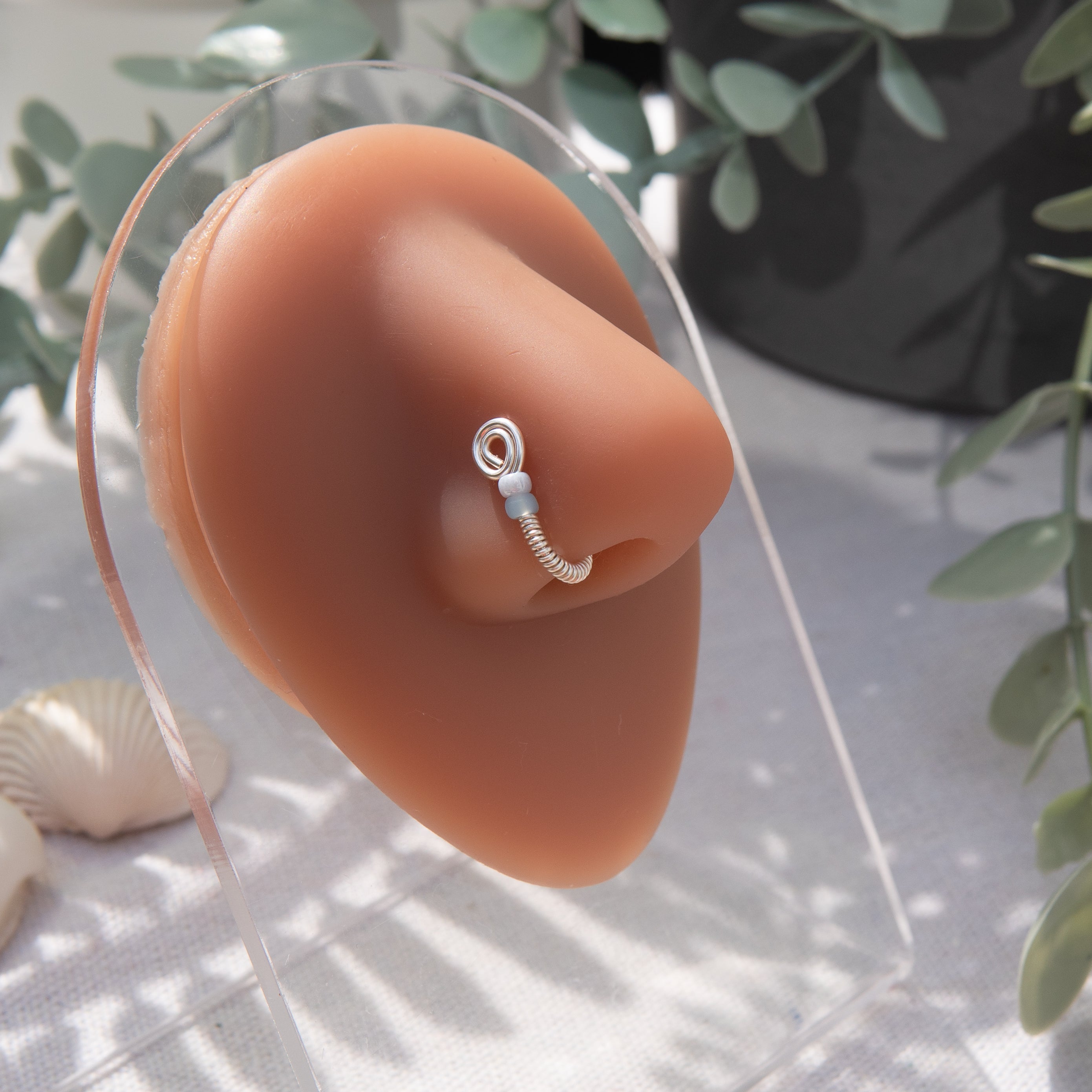 A silver-toned fake nose ring cuffs the model's nostril. The wire design forms a circular shape. Two seed beads rest below the circle. 