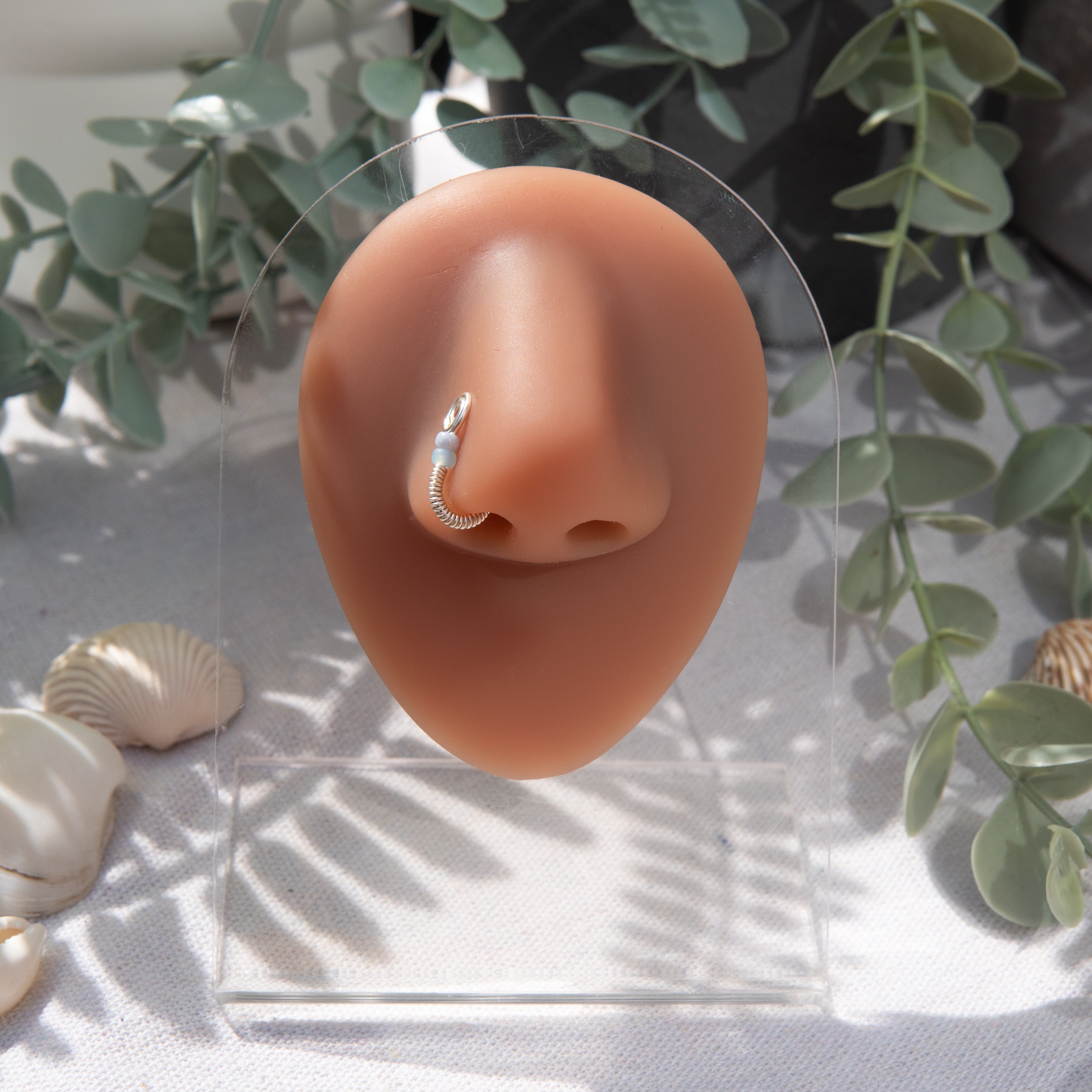 A silver-toned fake nose ring cuffs the model's nostril. The wire design forms a circular shape. Two seed beads rest below the circle. 